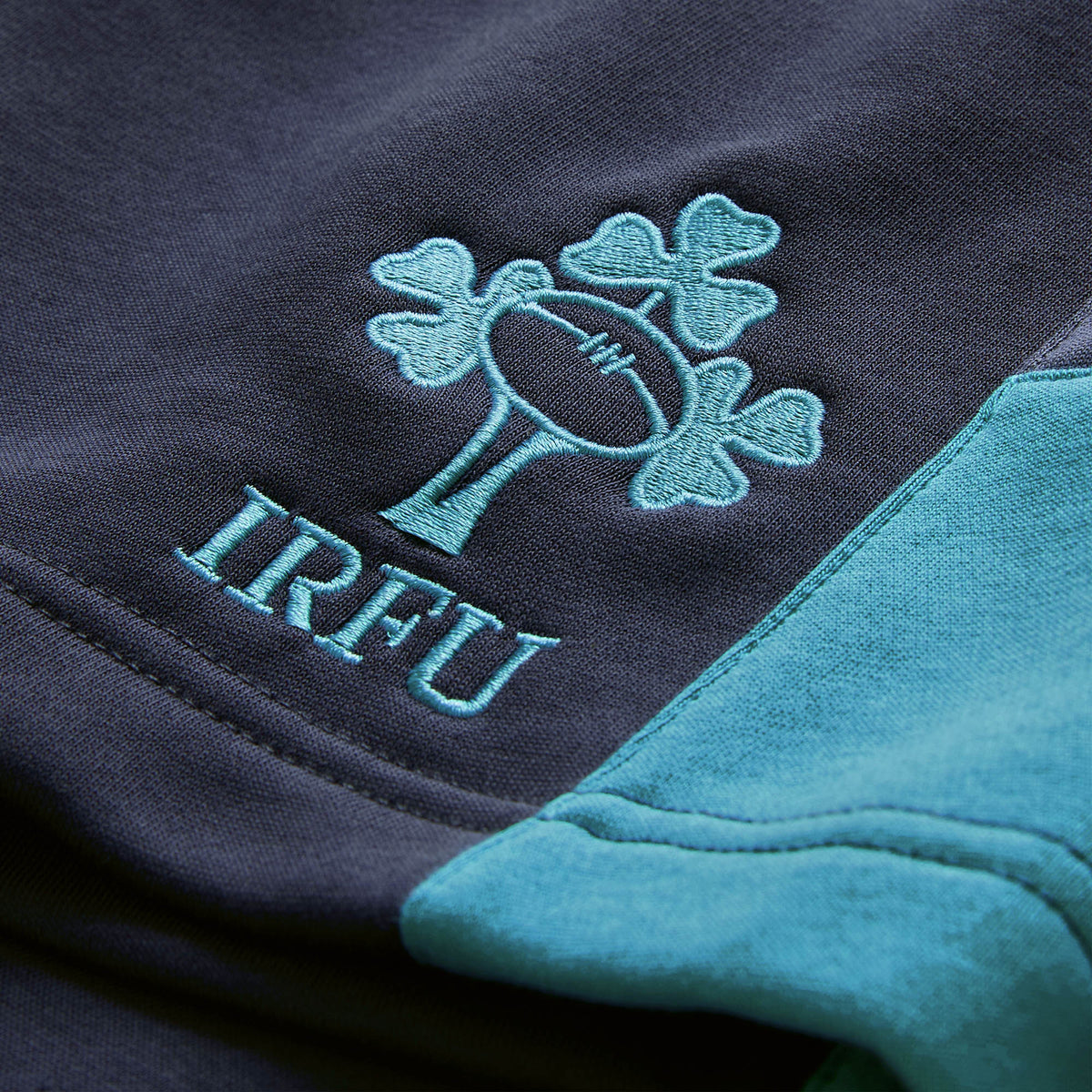 Close-up of the embroidered IRFU logo with a rugby ball and shamrocks on dark fabric, set beside lighter teal-colored material—highlighting the iconic detailing of the Ireland 26 Legends 7″ Shorts by Canterbury.