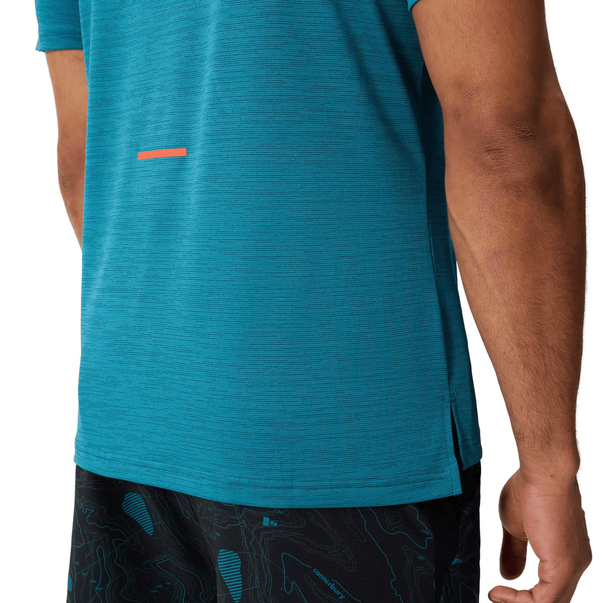 A person is shown from the side wearing a teal Canterbury Tempo Relaxed T-Shirt by Canterbury, made from breathable fabric, and black patterned shorts with their arm visible.