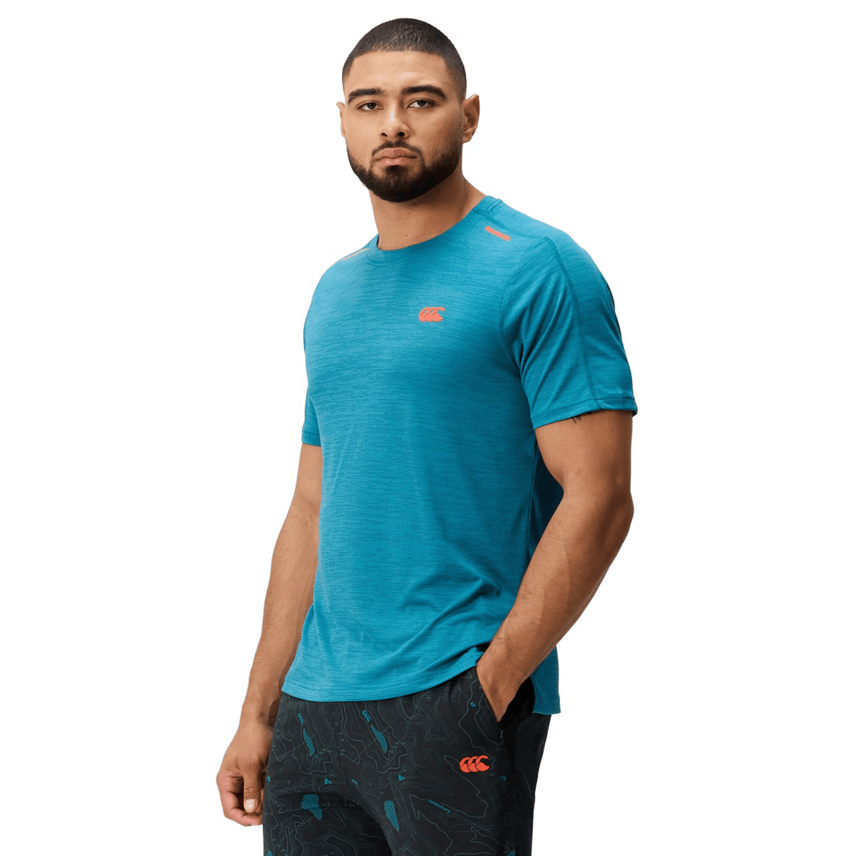 A man with short hair and a beard wears the Canterbury Tempo Relaxed T-Shirt by Canterbury and patterned shorts, standing in front of a plain white background.