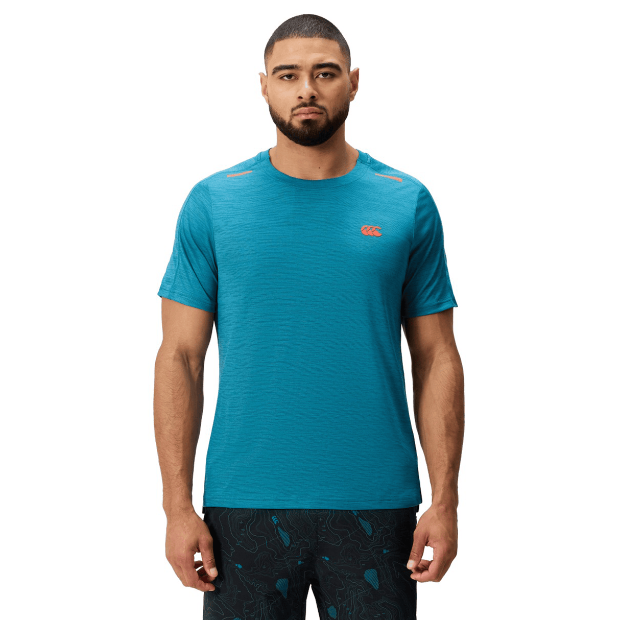 A man faces forward wearing a turquoise Canterbury Tempo Relaxed T-Shirt by Canterbury, made from breathable fabric, featuring a small red logo and paired with black patterned shorts.