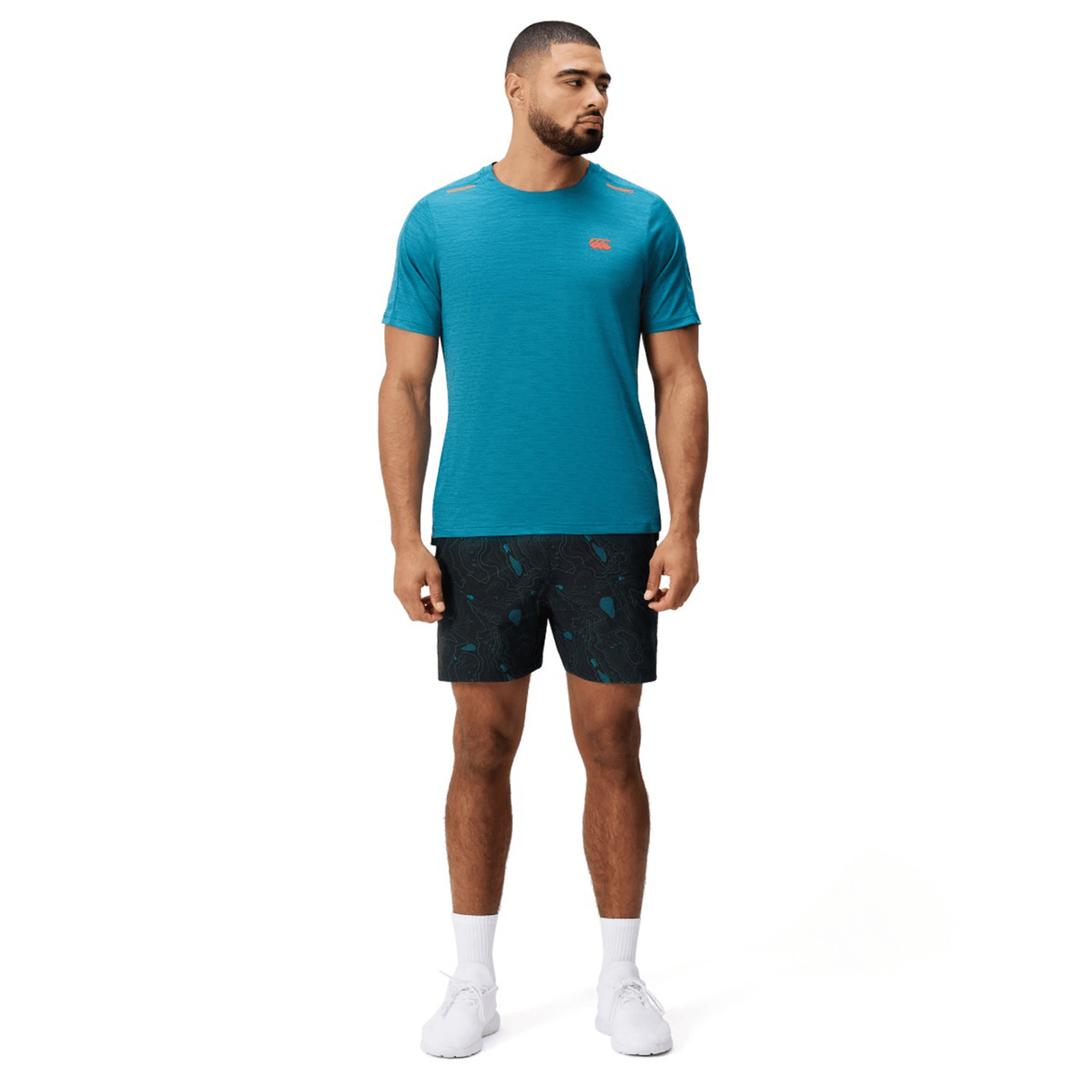A man faces forward on a white background, wearing the Canterbury Tempo Relaxed T-Shirt in teal by Canterbury, paired with black patterned shorts, white socks, and white sneakers.