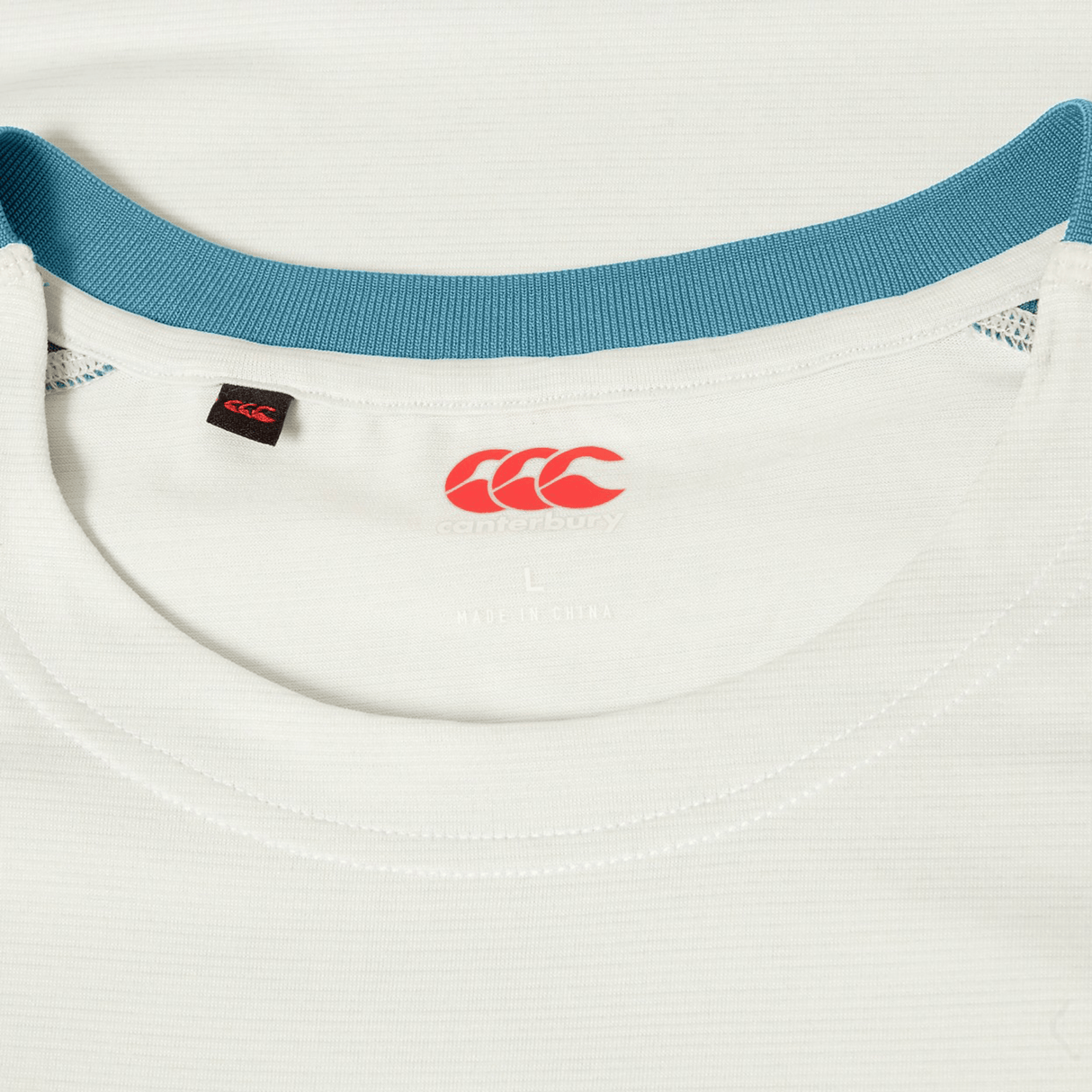 Close-up of the Canterbury Tempo Relaxed T-Shirt in white with a teal collar, featuring Canterbury’s red logo and interior size label. Designed with breathable fabric for comfort and performance.