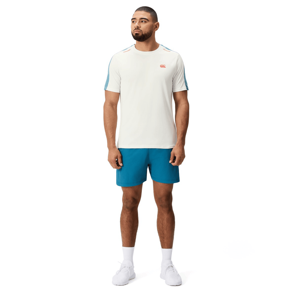 A man stands facing forward in a Canterbury Tempo Relaxed T-Shirt by Canterbury with blue accents, paired with blue shorts, white socks, and white sneakers against a plain background.