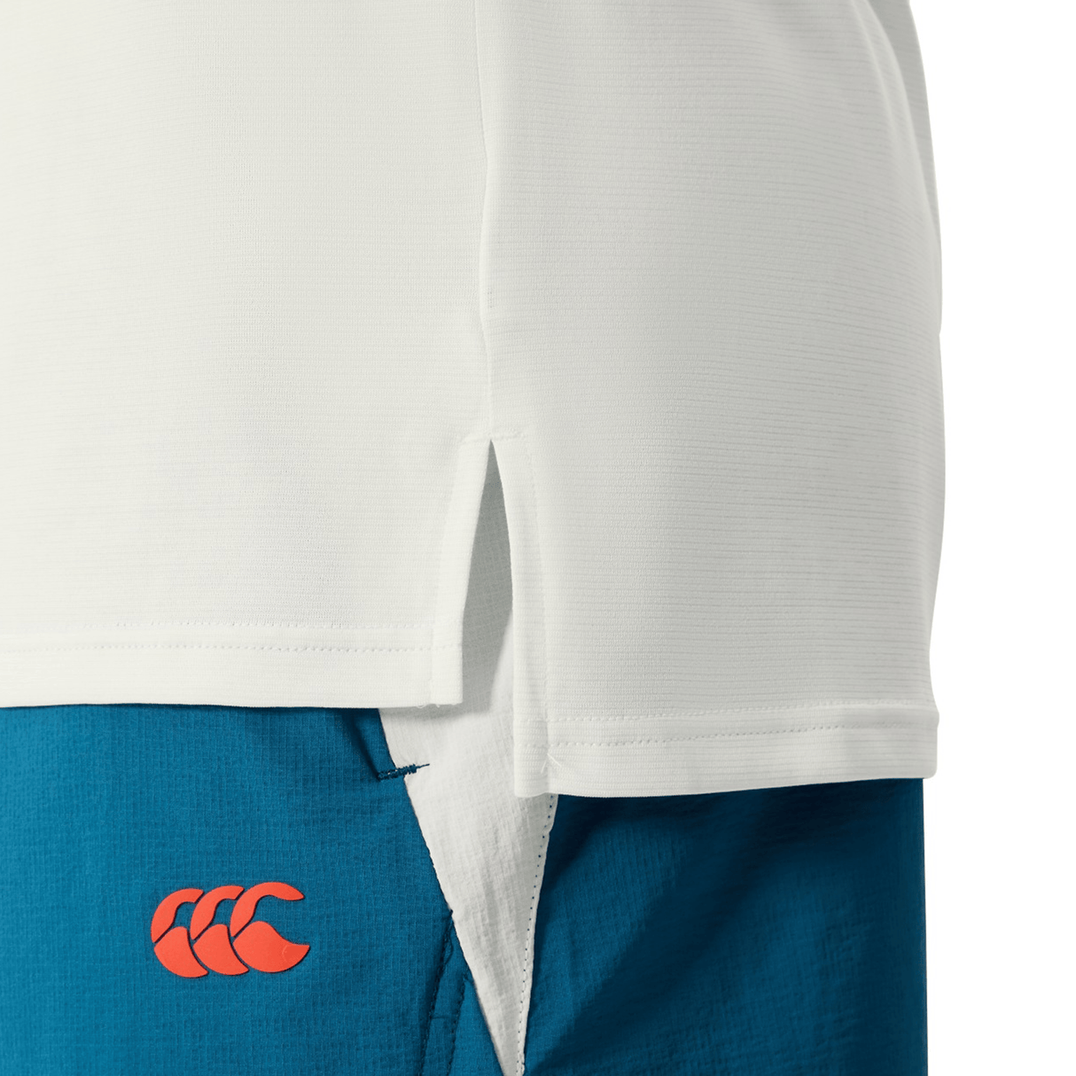Close-up of the Canterbury Tempo Relaxed T-Shirt, crafted from breathable fabric with a side slit, layered over blue shorts with a red Canterbury logo on the left side—ideal for a performance-ready look.