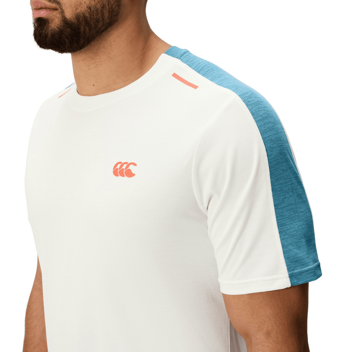 A person is shown from the chest up wearing the Canterbury Tempo Relaxed T-Shirt in white, featuring blue shoulder panels, a small coral chest logo, and breathable fabric. The background is plain.