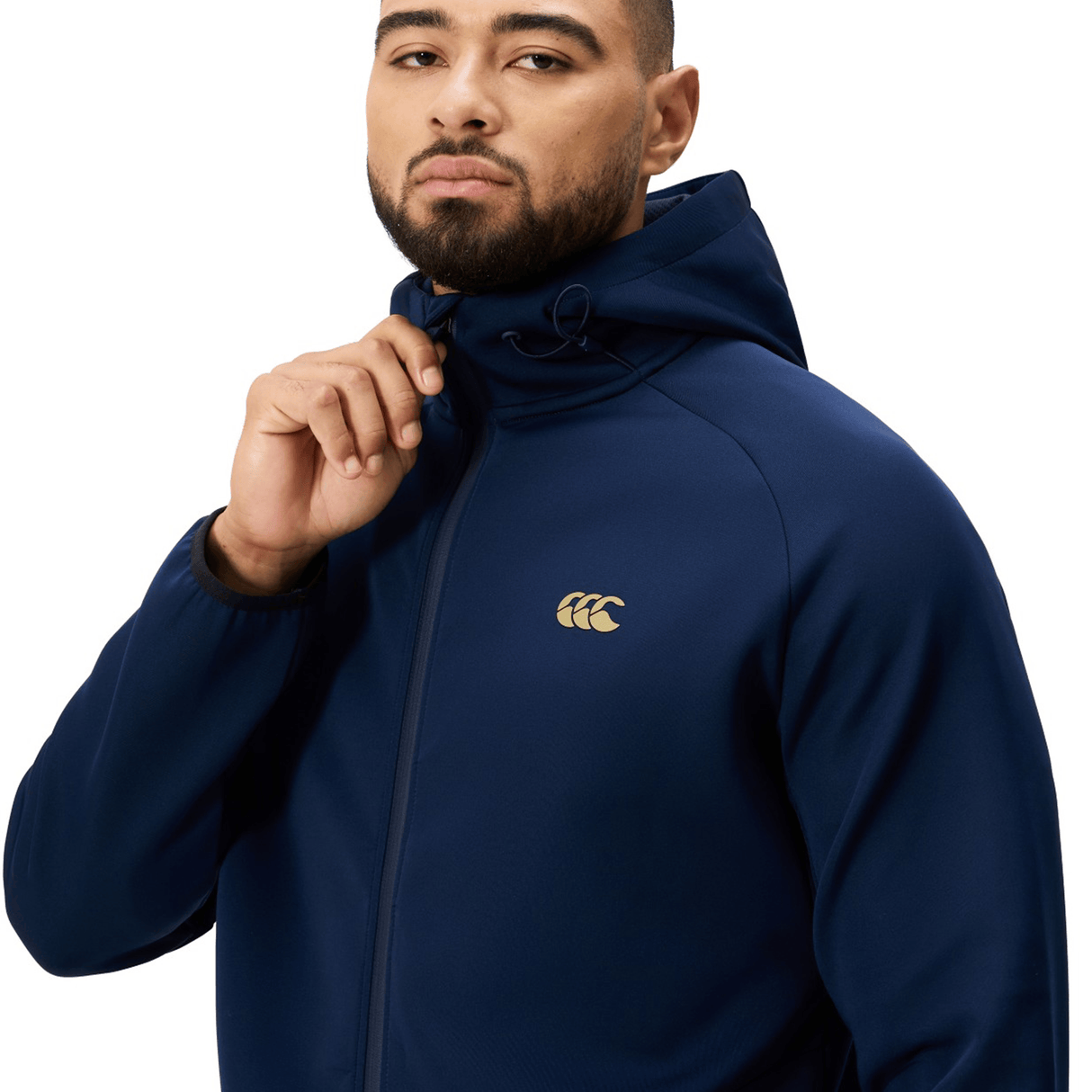 A man wears the Canterbury Everest Full Zip Hoodie in navy with a gold logo on the chest, holding the hood strings and looking at the camera against a white background. This Canterbury hoodie is ideal for workouts or casual wear.