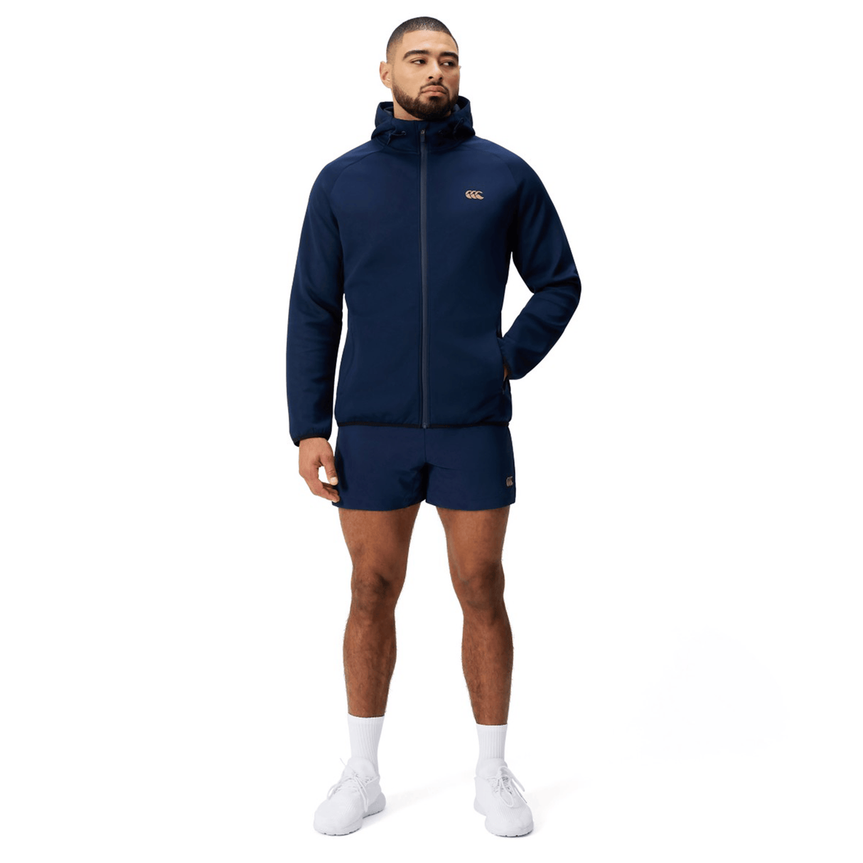 A man faces forward wearing a navy blue Canterbury Everest Full Zip Hoodie and matching shorts, paired with white socks and sneakers, set against a plain white background.