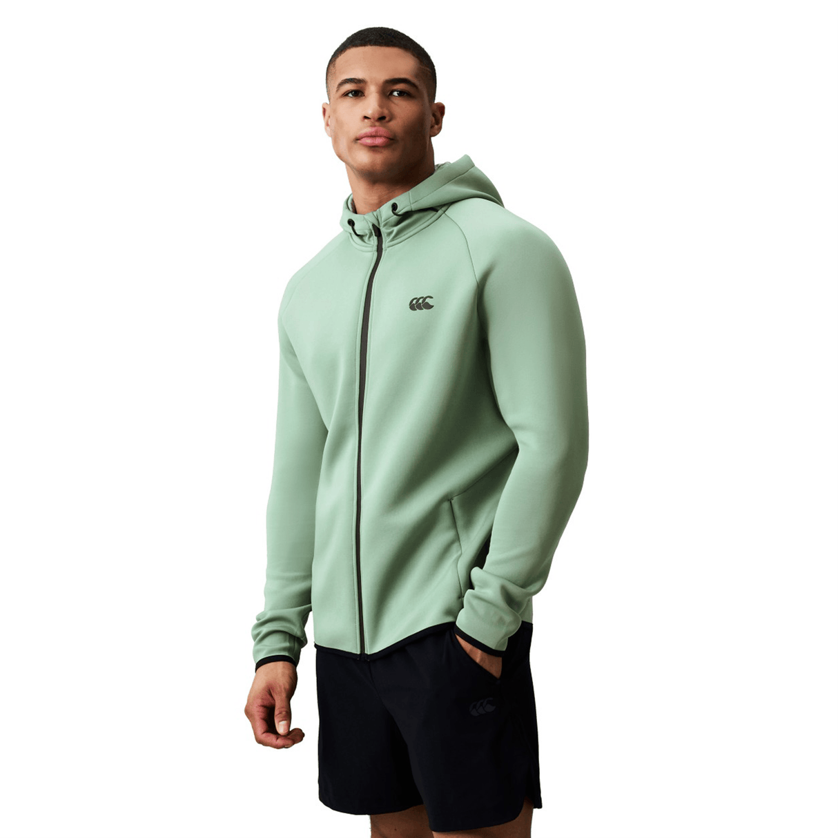 A man wears a light green Canterbury Everest Zip Thru Hoodie by Canterbury, featuring a hood, paired with black shorts, standing against a plain white background.