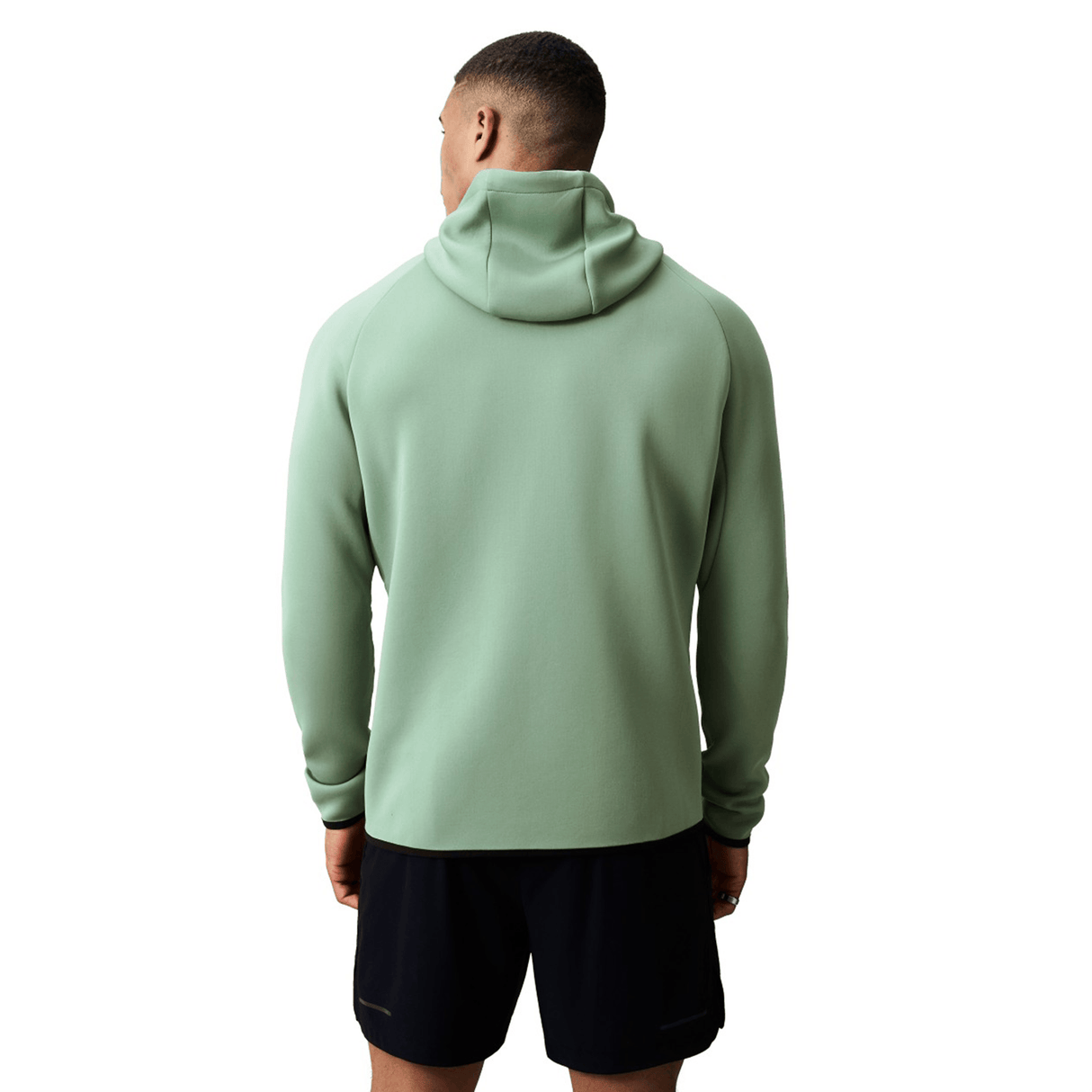 A person wearing a light green Canterbury Everest Zip Thru Hoodie by Canterbury and black shorts stands with their back to the camera against a white background.