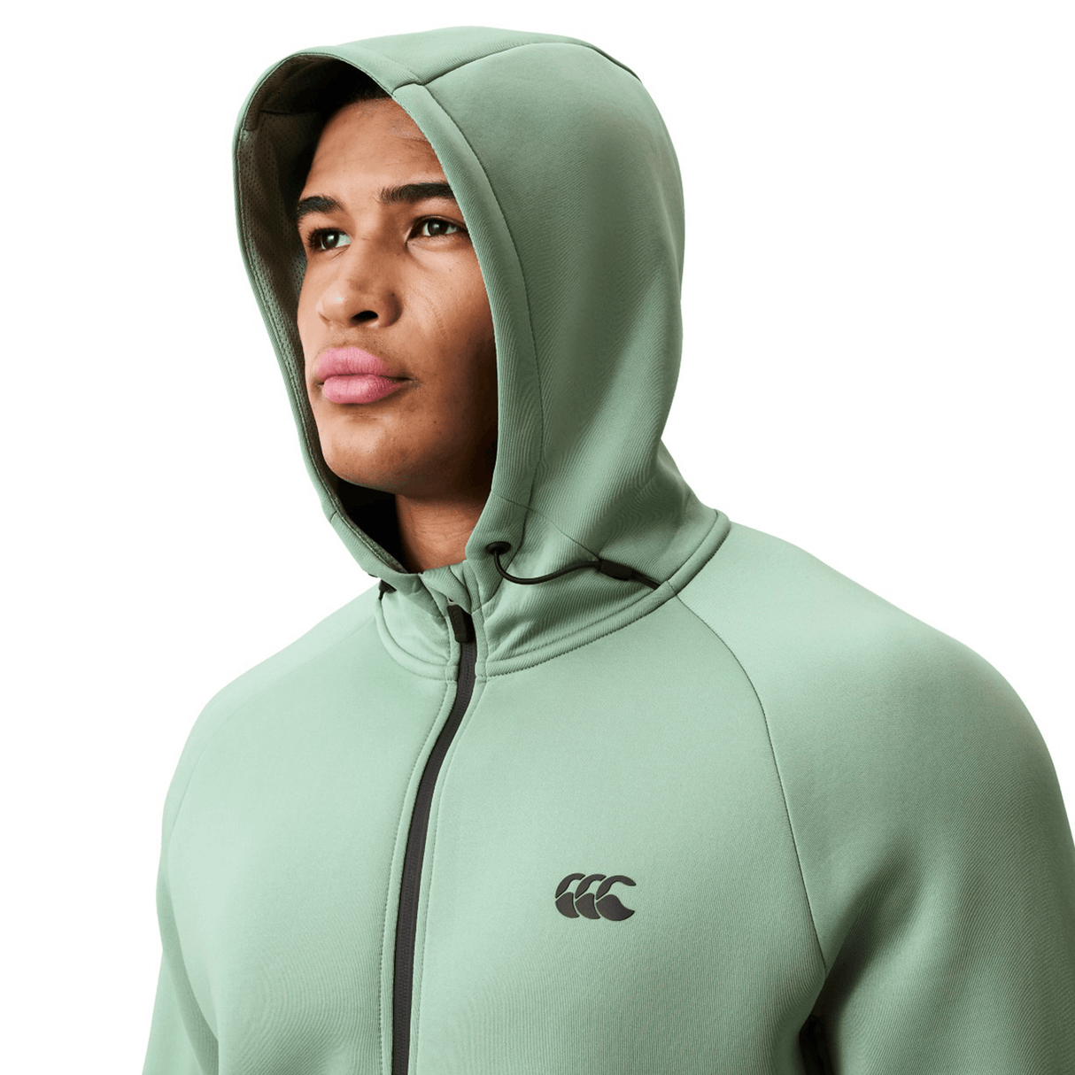 A person wears a light green Canterbury Everest Zip Thru Hoodie by Canterbury, hood up, featuring a black logo on the chest, looking slightly upward.