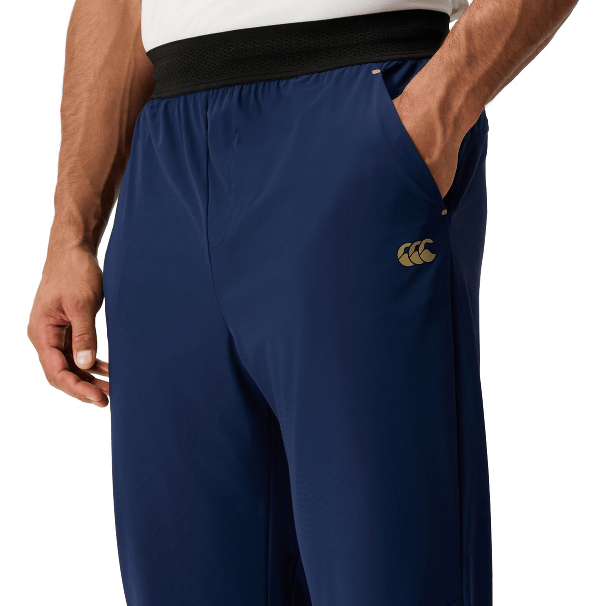 Someone wearing Canterbury Everest Woven Pant 30" in navy with a gold thigh logo stands with one hand in a pocket, highlighting the sweat-wicking comfort.