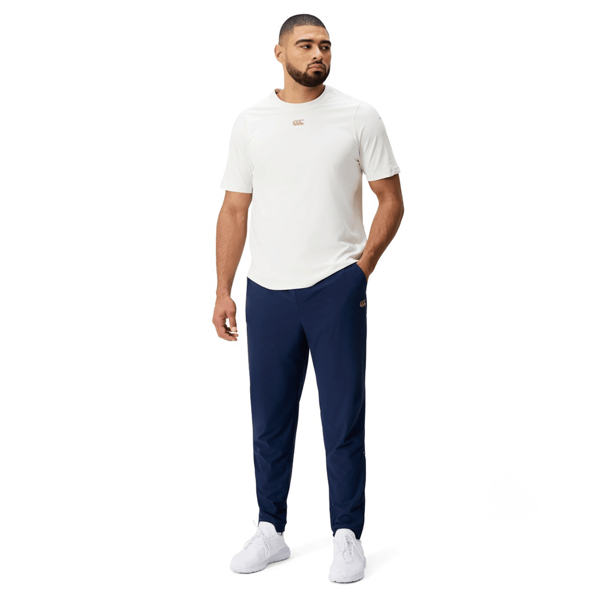 A man faces slightly left, wearing a white short-sleeve shirt, Canterbury Everest Woven Pant 30" by Canterbury in navy blue, and white sneakers against a plain white background.