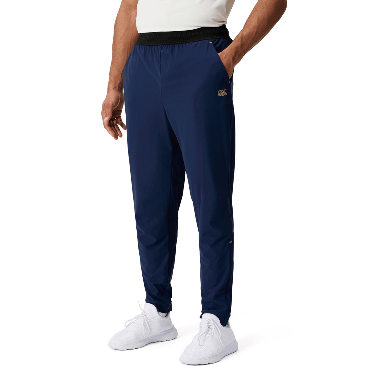 A person wears Canterbury Everest Woven Pant 30" in navy with a black waistband and four-way stretch, paired with white shoes and a white shirt, standing against a plain white background.