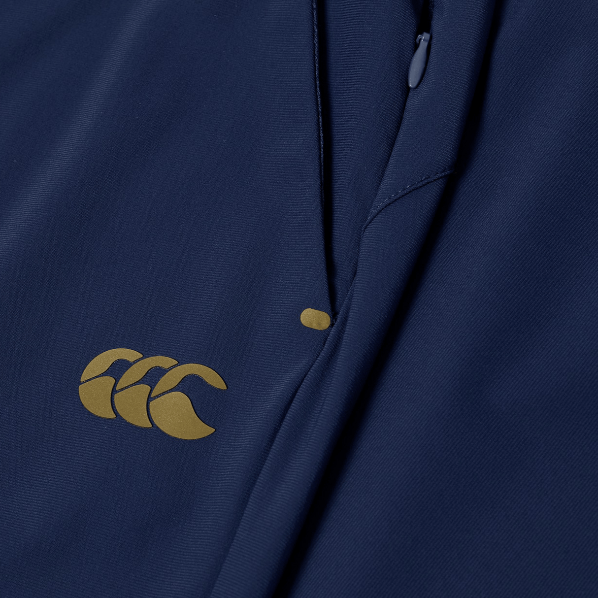 Close-up of the Canterbury Everest Woven Pant 30" in navy blue, featuring a yellow Canterbury logo, sweat-wicking fabric, and a partially visible zipper.
