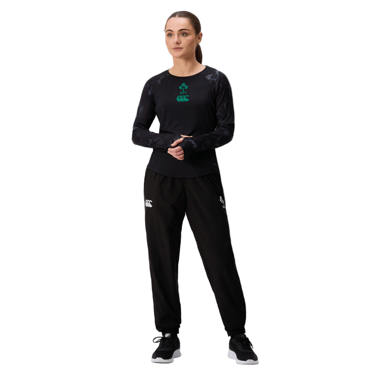 A woman stands against a white background wearing a black long-sleeve shirt, black sneakers, and Canterbury Women's Ireland 25/26 Training Pants featuring branded logos.