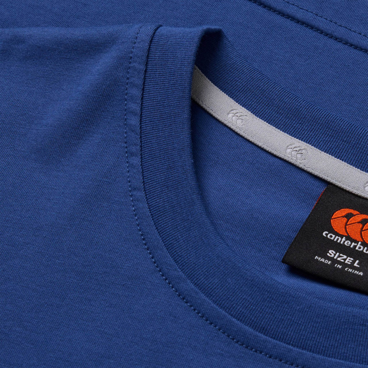 Close-up of two blue Canterbury Fundamentals T-Shirts, one partially overlapping the other. The visible tag displays the Canterbury brand and size L, with “Made in China,” making these Canterbury Fundamentals T-Shirts a versatile choice for athletic casual wear or as a classic rugby t-shirt.