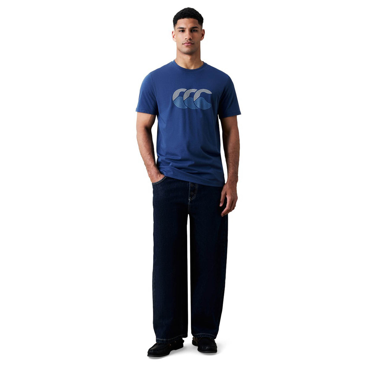 A man stands facing forward against a plain white background, wearing the Canterbury Fundamentals T-shirt by Canterbury styled as athletic casual wear, paired with dark wide-leg jeans and black shoes.