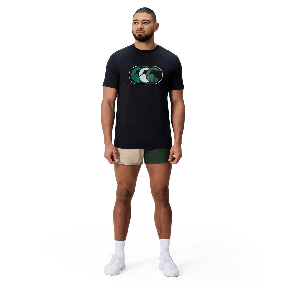 A man stands on a white background wearing a Canterbury FUN. T-SHIRT AM in black with a green wave logo, paired with beige and green two-tone shorts, white socks, and white sneakers.
