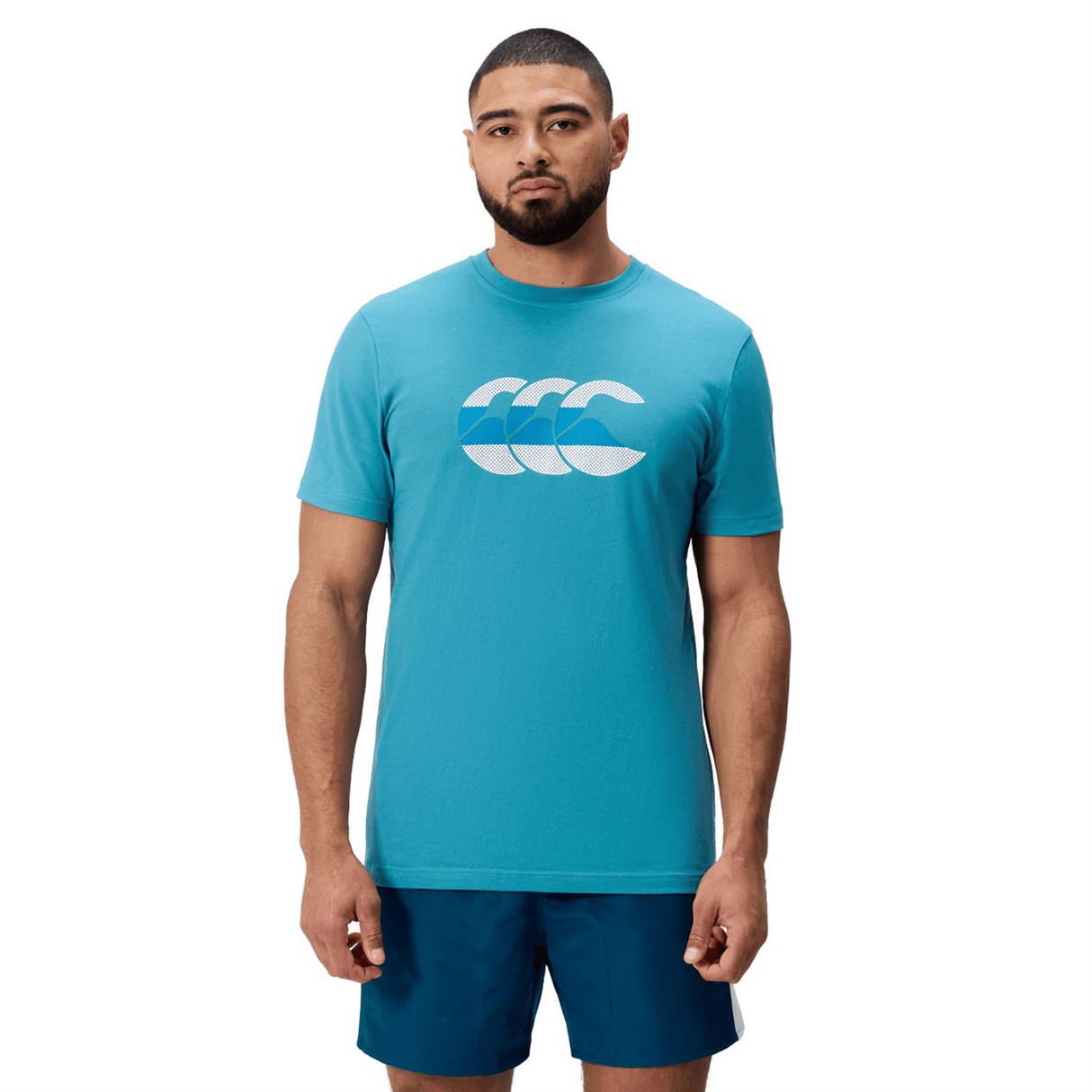 A man faces forward wearing a Canterbury FUN. T-SHIRT AM in turquoise with a white and blue chest graphic, paired with dark blue shorts, set against a white background.