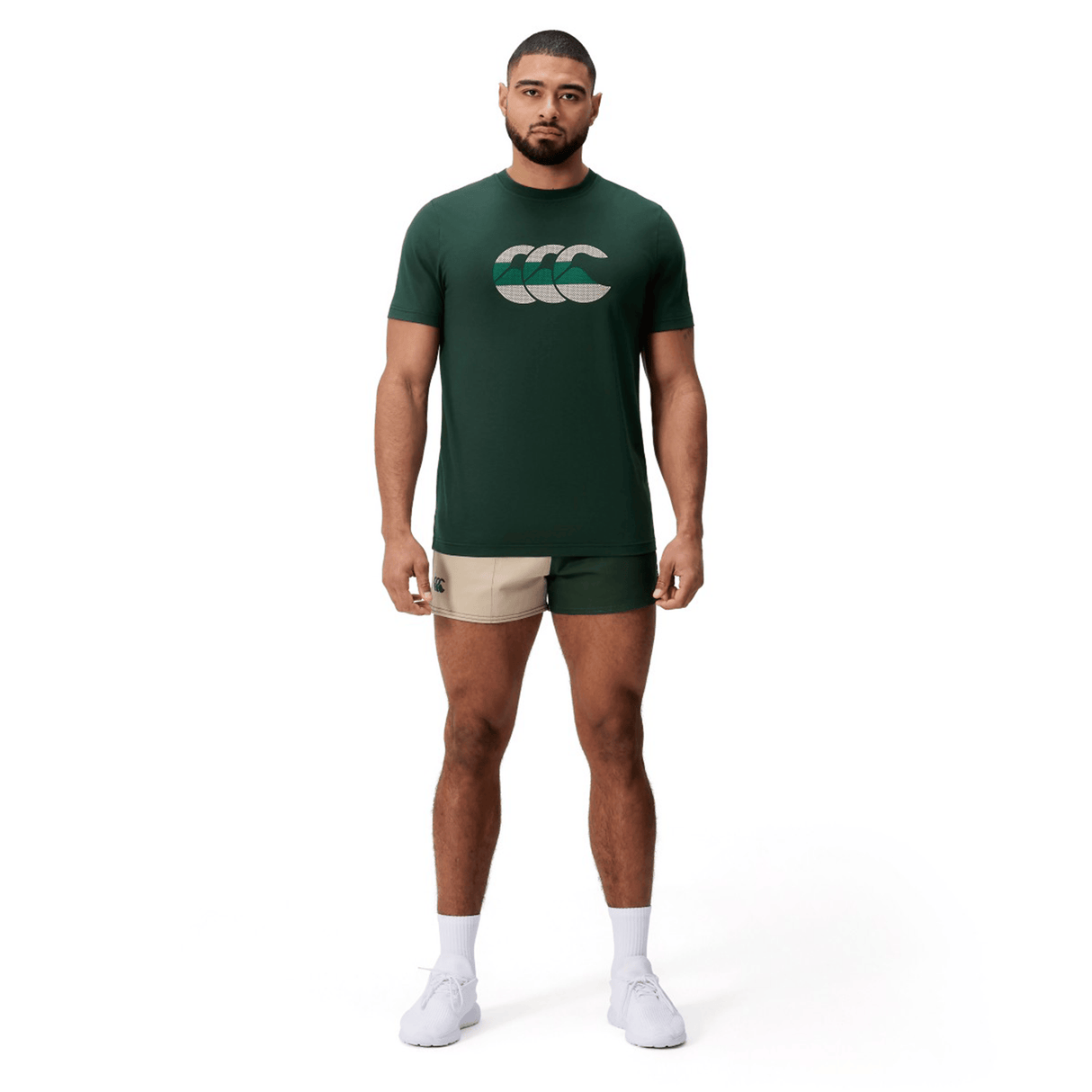A man faces forward against a white background, wearing a dark green Canterbury FUN. T-SHIRT AM with logo, beige athletic shorts, white socks, and white shoes.
