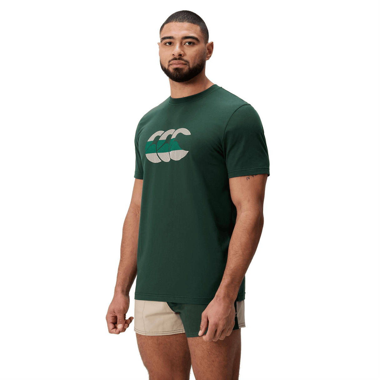 A man stands at an angle against a white background, wearing a Canterbury FUN. T-SHIRT AM in dark green with a bold logo and beige-green shorts.
