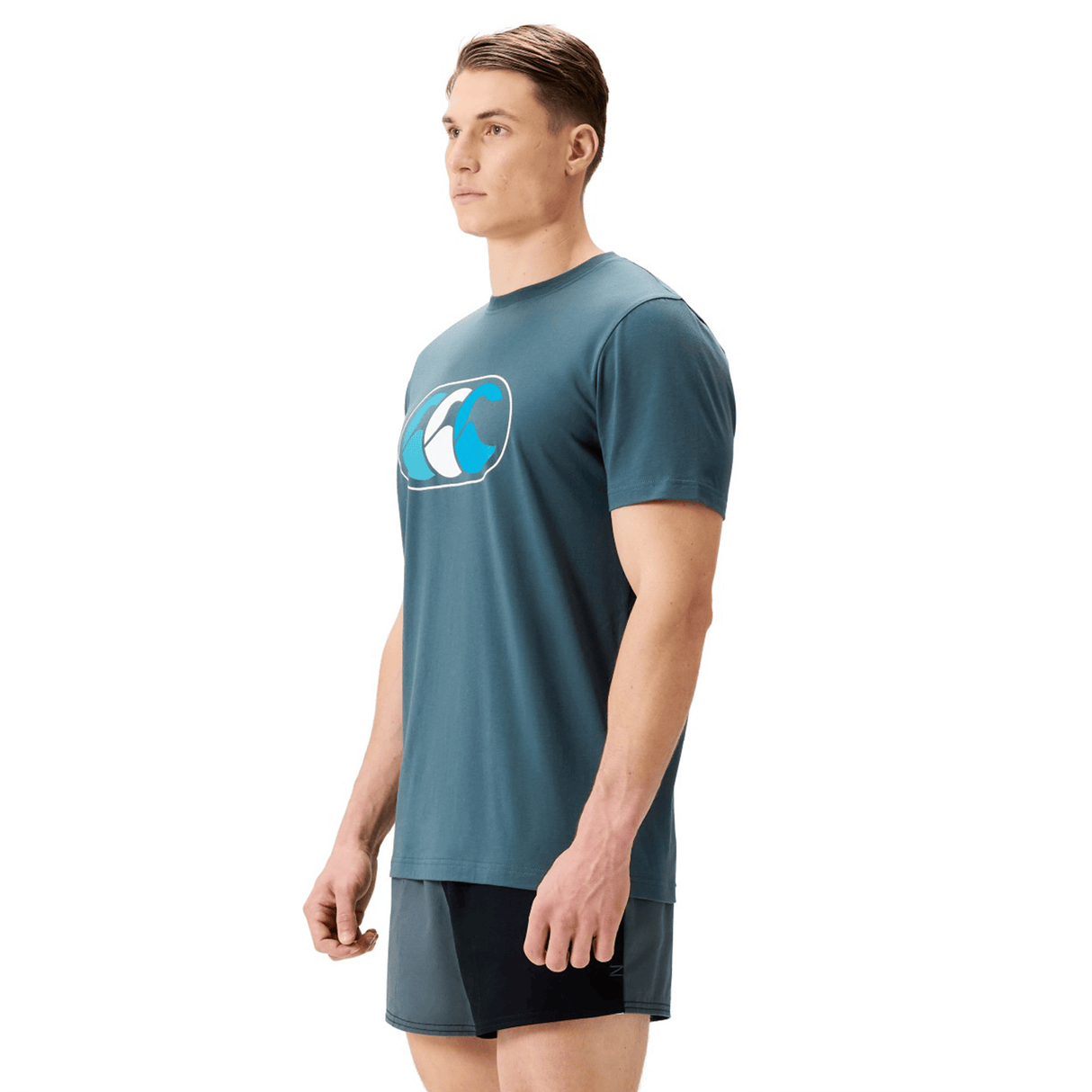 A man wearing the Canterbury FUN. T-SHIRT AM, a teal graphic t-shirt with a wave logo, and black athletic shorts stands facing left against a plain white background.