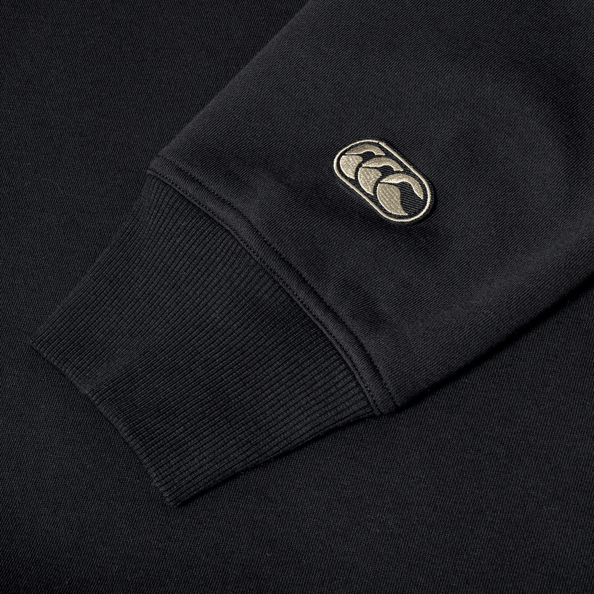 Canterbury's EVERYDAY GFX CREW SWEATER features black sweatshirt sleeves with ribbed cuffs and an embroidered beige and black logo patch near the wrist—ideal for daily wear or pairing with your favorite sweater.