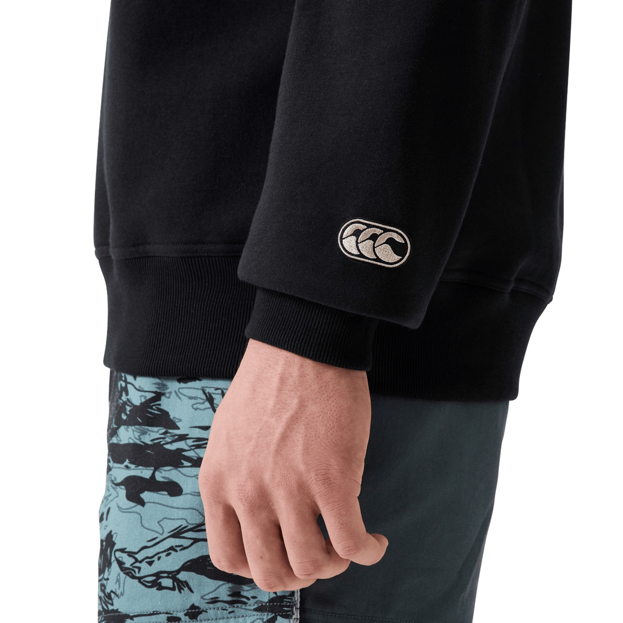 A person wears a black Canterbury EVERYDAY GFX CREW SWEATER with a white sleeve logo and teal shorts featuring a black abstract pattern.