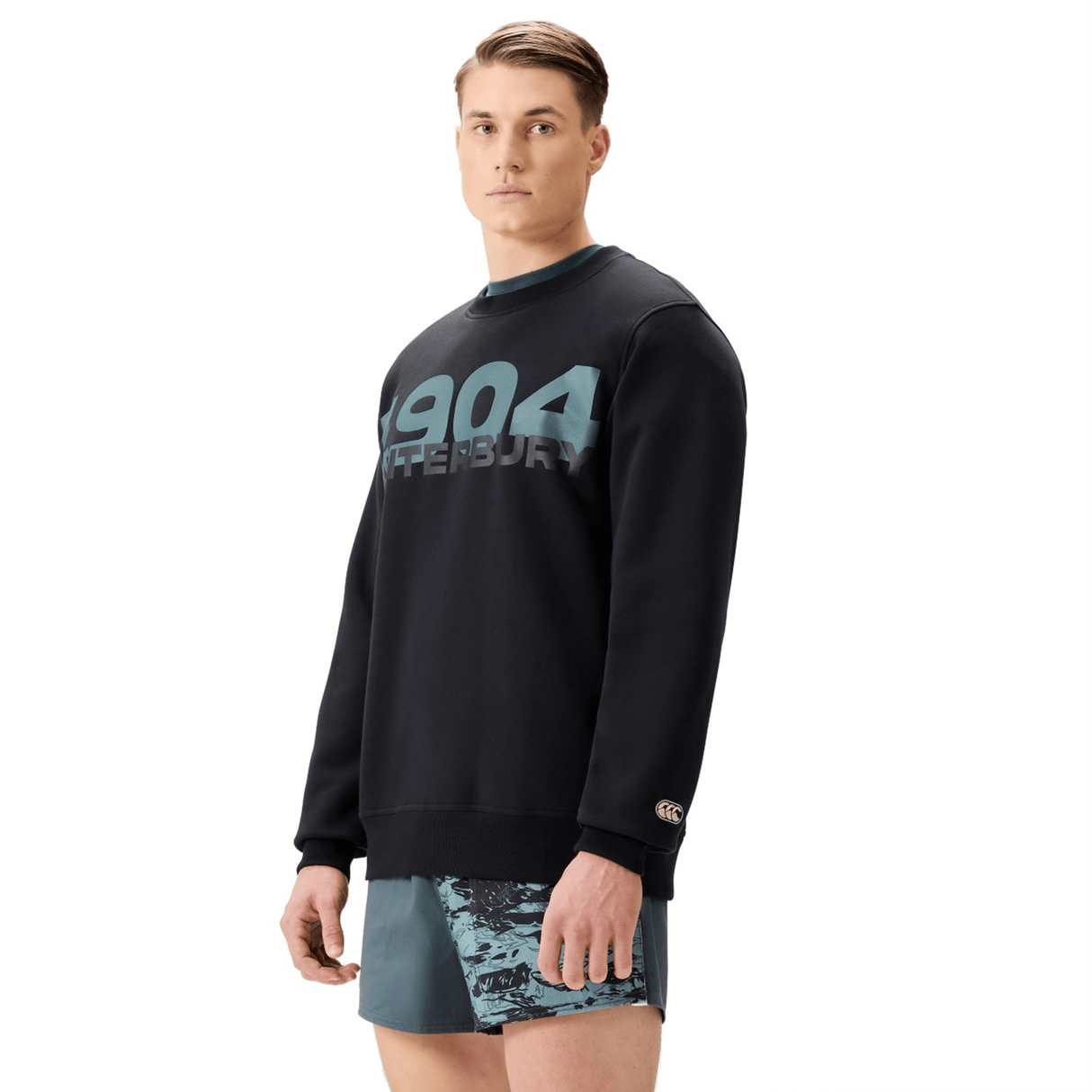 A man stands facing forward in a black Canterbury EVERYDAY GFX CREW SWEATER with "1904 Canterbury" on the front and patterned athletic shorts, showcasing a classic everyday look from Canterbury.