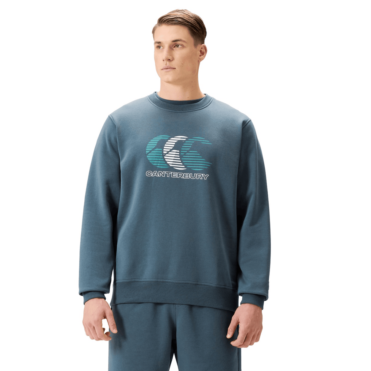A man models the Canterbury EVERYDAY GFX CREW SWEATER in teal, paired with matching pants, standing against a plain white background to showcase casual everyday style.