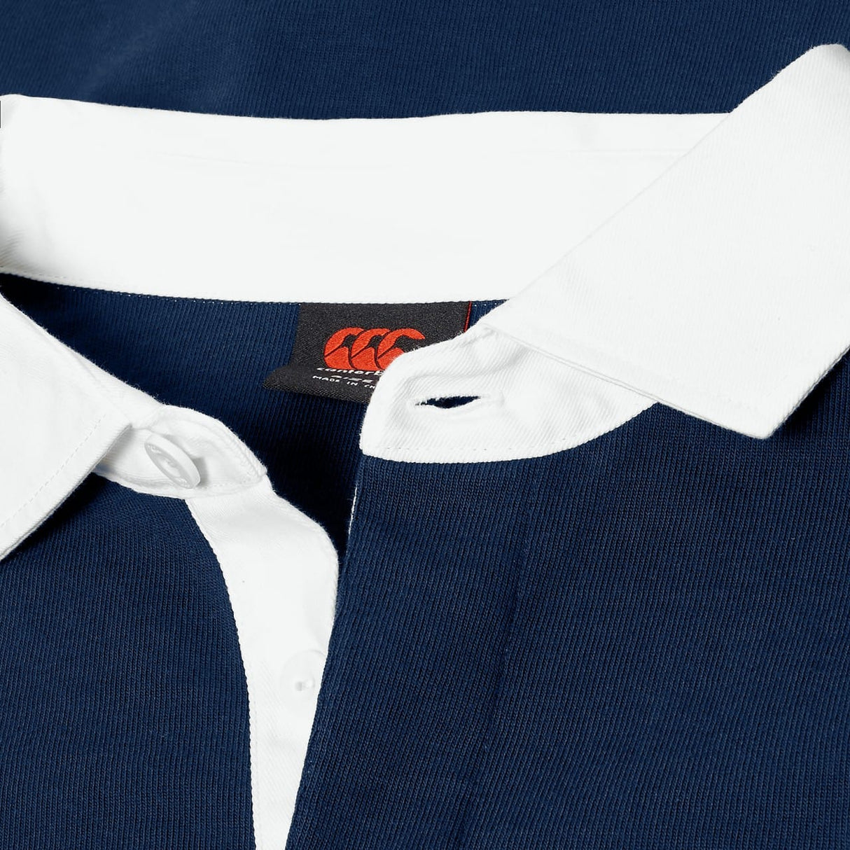 Close-up of the Canterbury Stripe Rugby Jersey by Canterbury, featuring a navy blue color, white collar and placket with visible buttons, and a black tag with red logo inside the collar. Classic rugby style inspired by traditional designs.