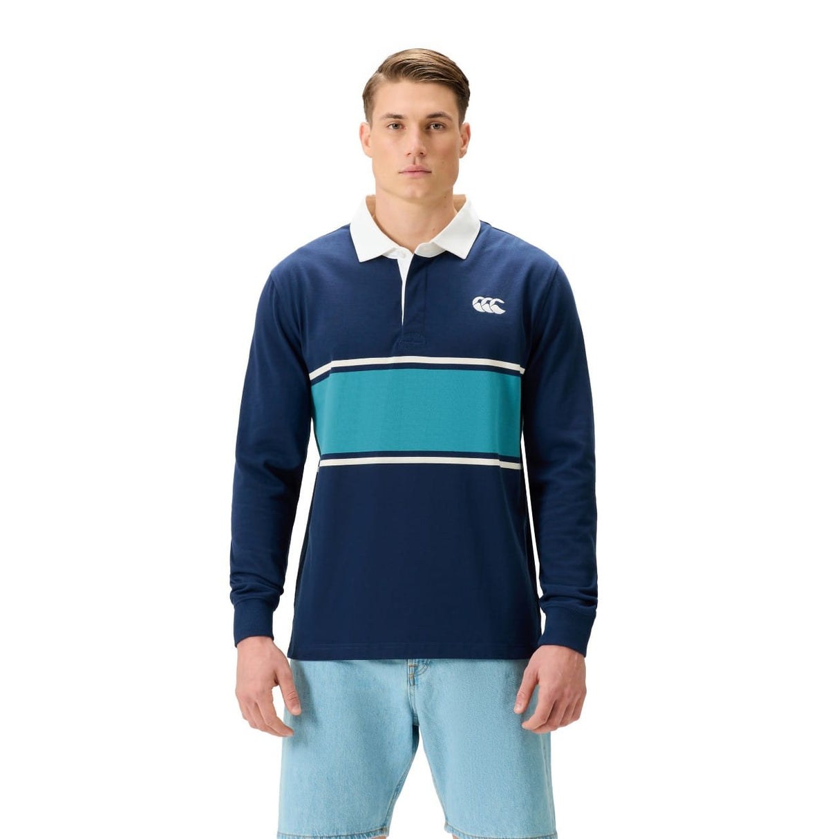 A man wearing a Canterbury Stripe Rugby Jersey by Canterbury, featuring navy blue with white and teal stripes and a white collar, paired with light blue shorts, stands facing forward against a white background.