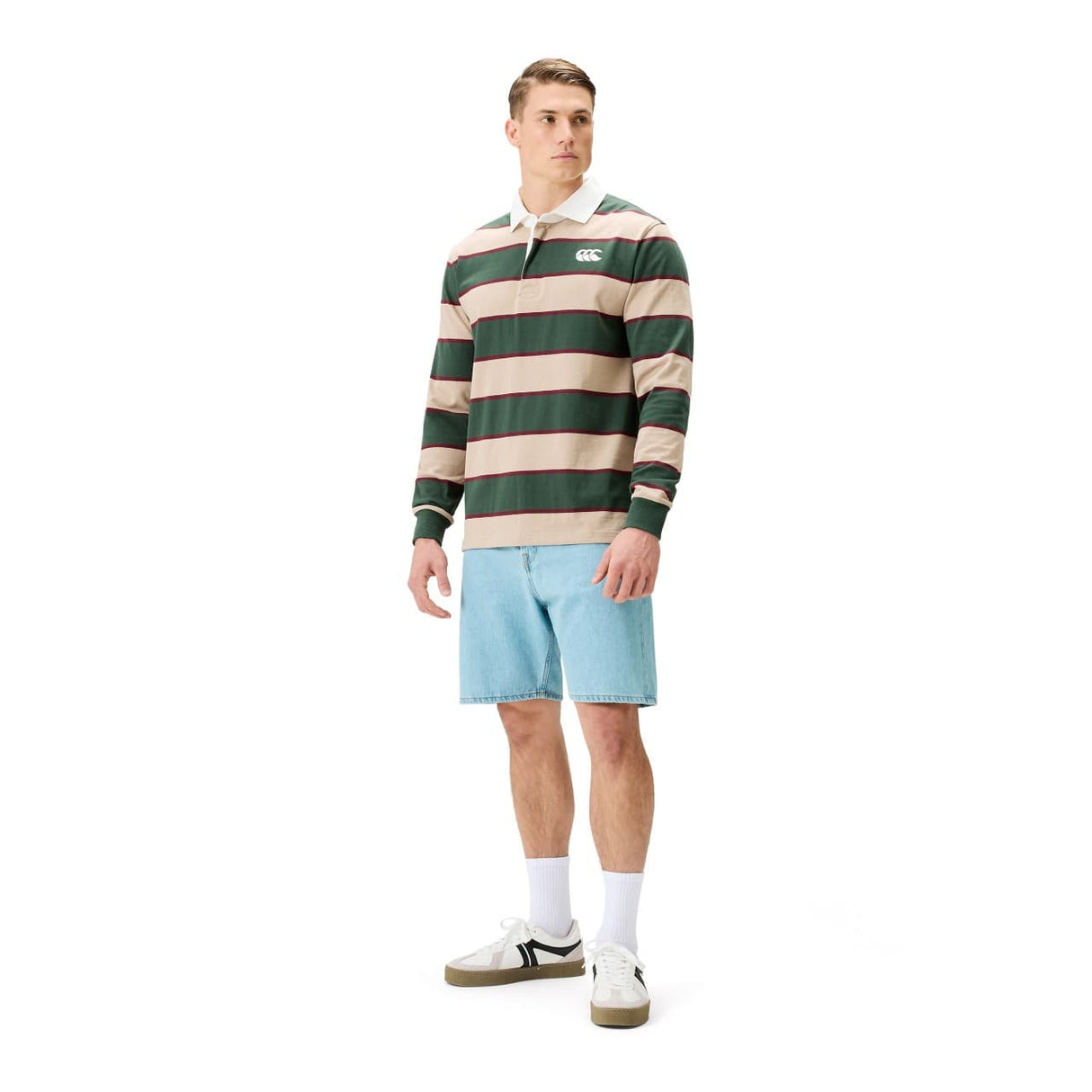 A man wearing a Canterbury Stripe Rugby Jersey by Canterbury in green and beige, paired with light blue denim shorts, white socks, and white sneakers stands slightly turned against a white background.
