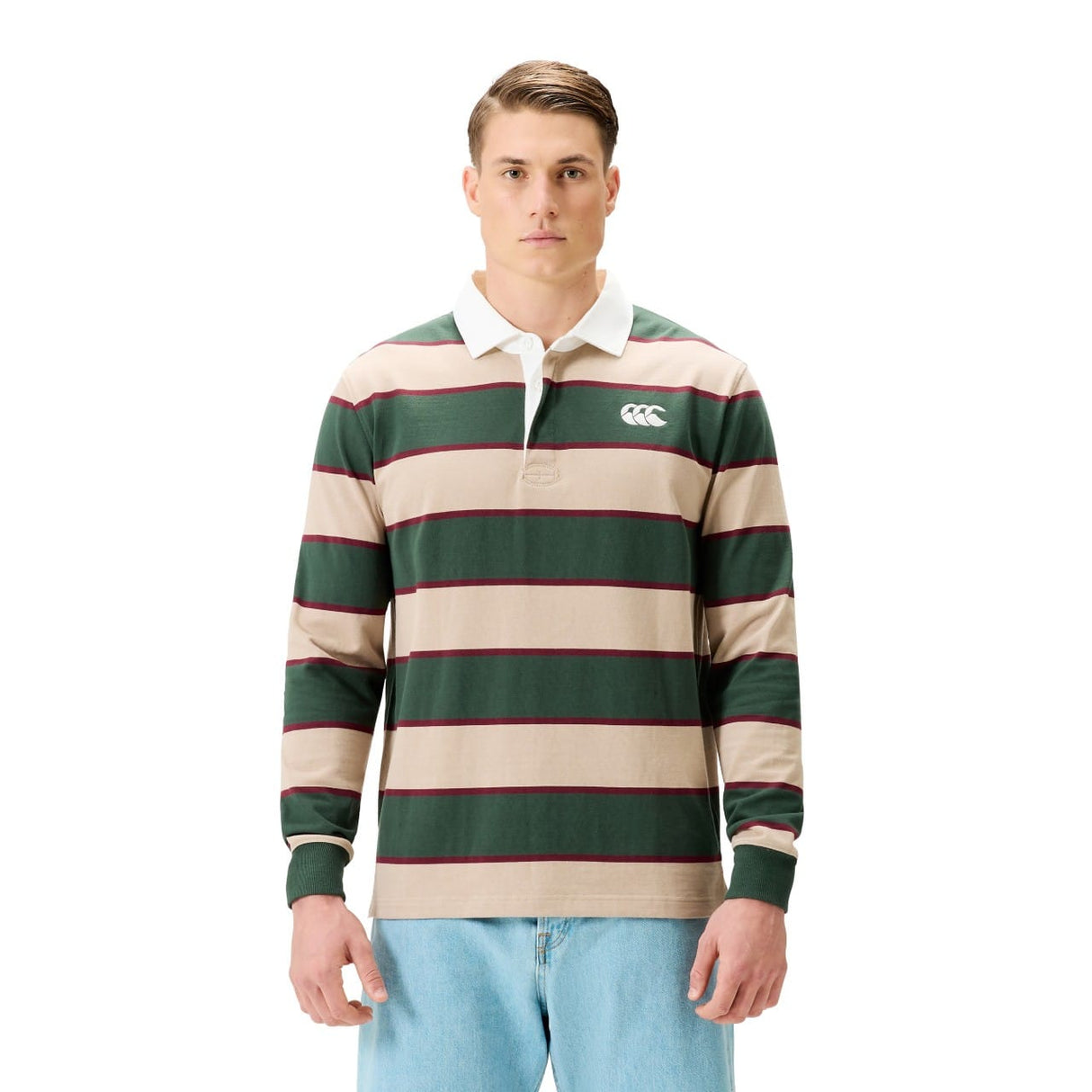 A man wears the Canterbury Stripe Rugby Jersey by Canterbury in green, beige, and red with a white collar, paired with blue jeans, standing against a plain white background.