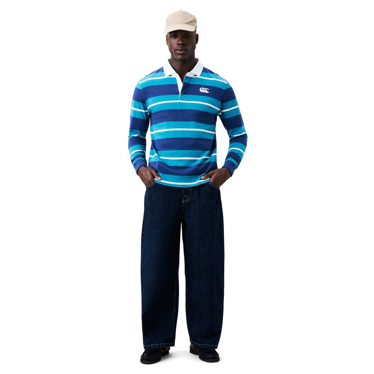 A man wears a cream cap, Canterbury Stripe Classic Rugby Jersey by Canterbury made from a durable cotton blend, wide-leg dark jeans, and black shoes, standing against a plain white background.