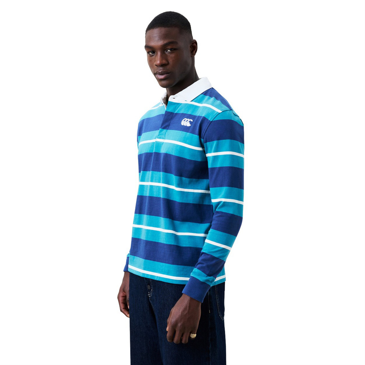 A man wears the Canterbury Stripe Classic Rugby Jersey by Canterbury, featuring blue and teal stripes, a white collar, and crafted from a durable cotton blend, standing against a plain white background.