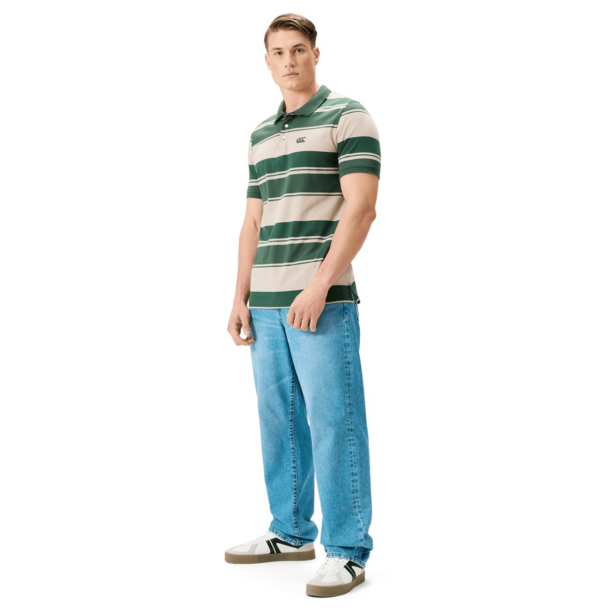 A man wears the Canterbury Yarn Stripe Polo by Canterbury in green and beige, paired with light blue jeans and white sneakers, capturing classic rugby style against a plain white background.