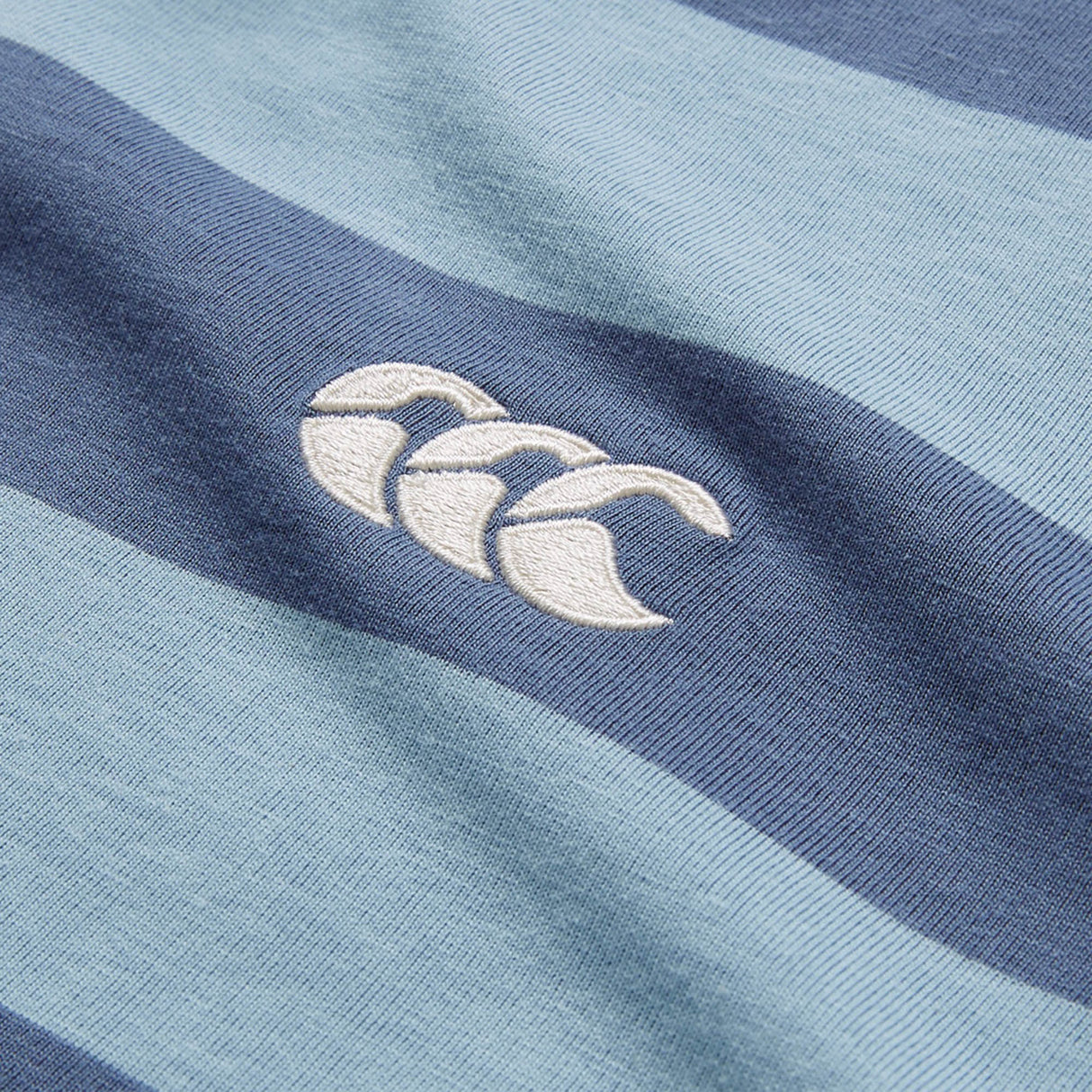 Close-up of the Canterbury Yarn Stripe Polo by Canterbury, featuring blue and navy rugby-style stripes and a white embroidered logo of three overlapping curved shapes.
