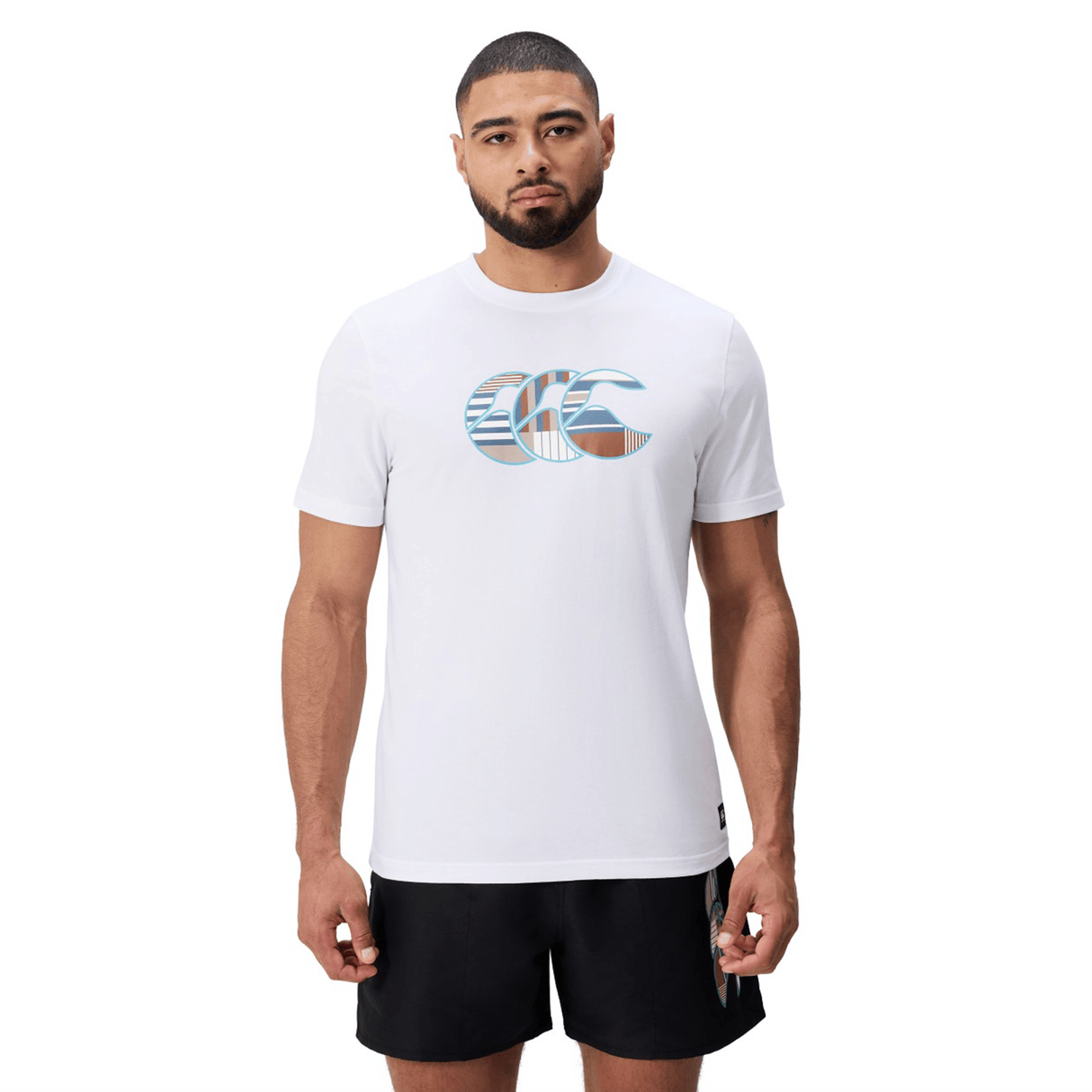 A man wearing a Canterbury Uglies Logo T-shirt, a white cotton blend tee with a multicolored graphic by Canterbury, and black shorts stands facing forward against a plain white background.