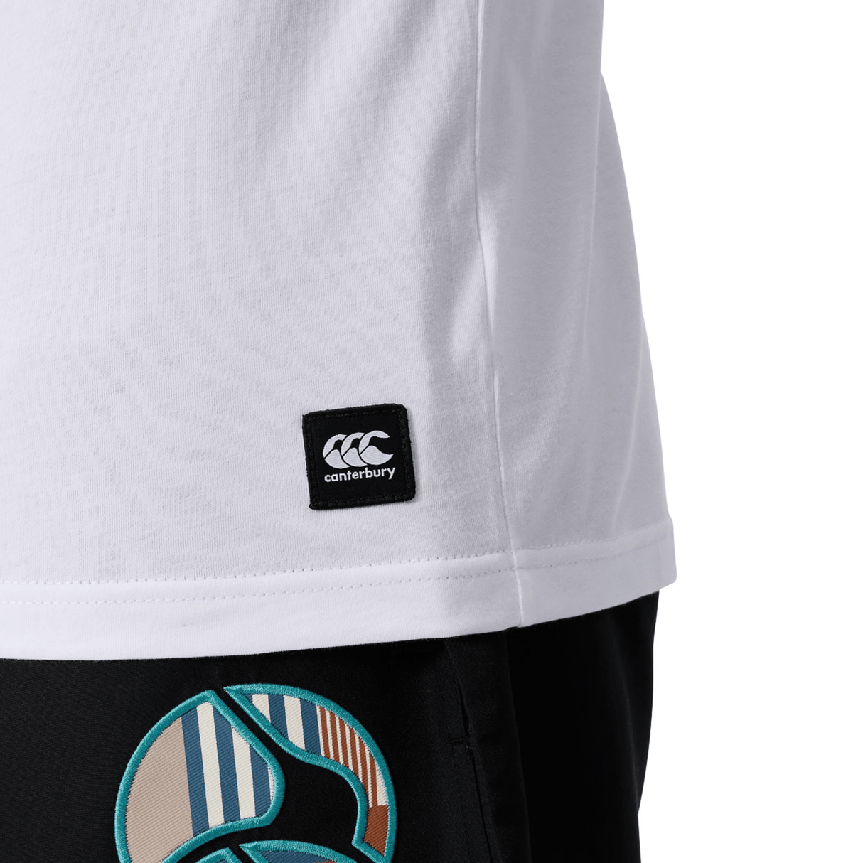 Close-up of a person in a white Canterbury Uglies Logo T-shirt by Canterbury, featuring a visible logo tag, paired with black pants with a colorful circular pattern.