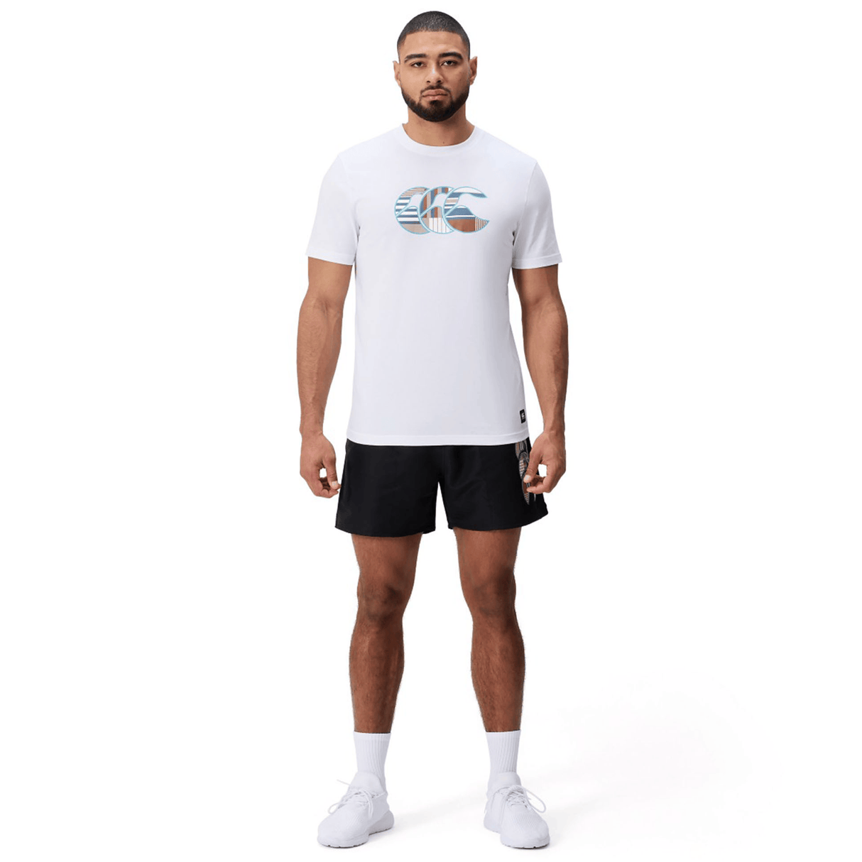 A man stands facing forward wearing a white Canterbury Uglies Logo T-shirt, black athletic shorts, and white sneakers against a plain white background.