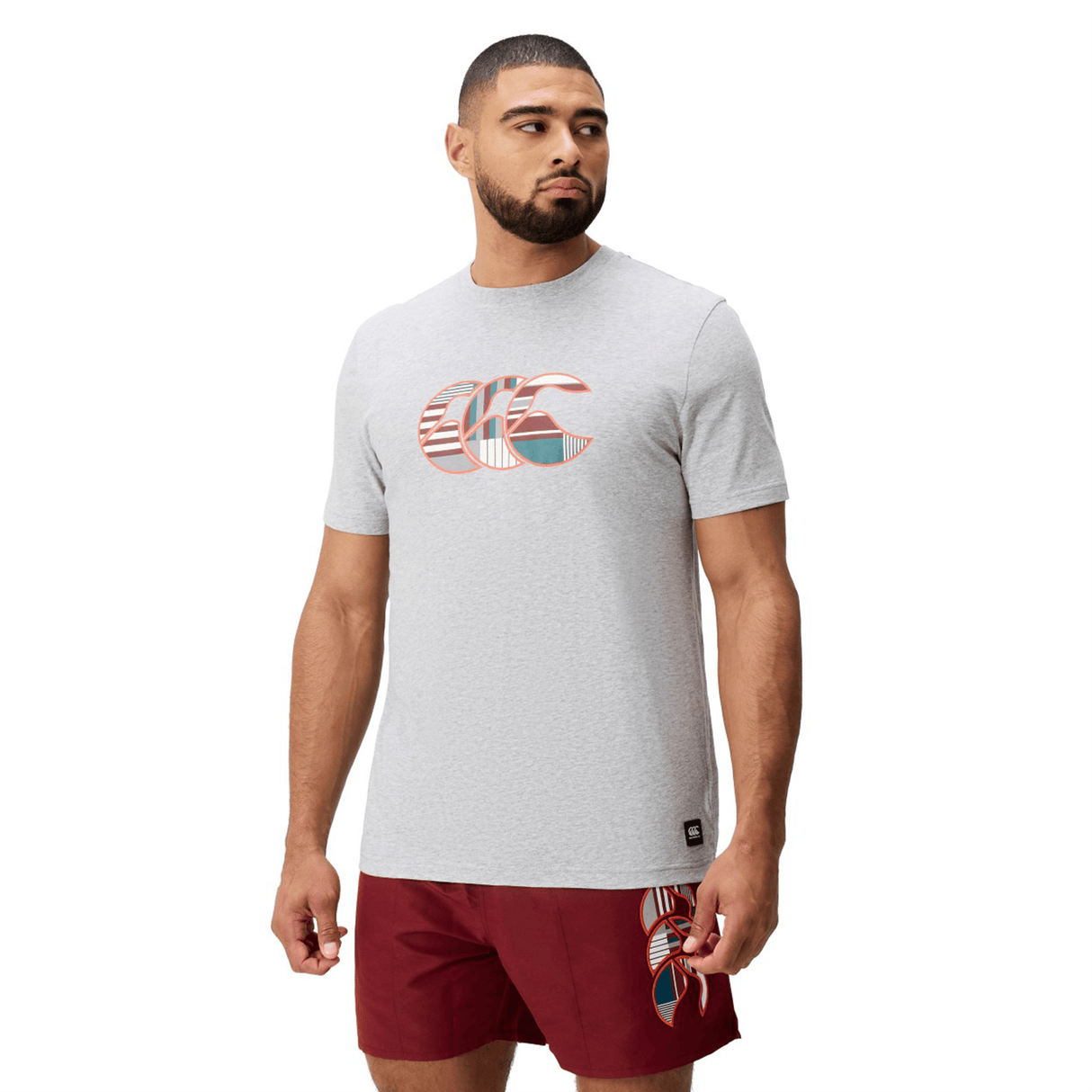 A man wearing a light gray Canterbury Uglies Logo T-shirt and red shorts stands turned slightly to the side against a plain white background.