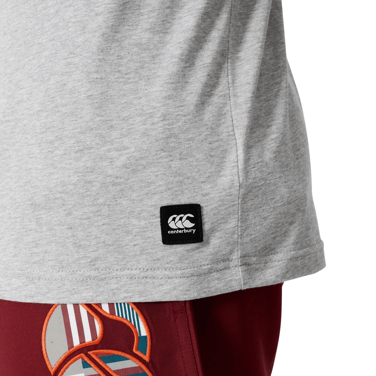 A close-up of a person wearing the Canterbury Uglies Logo T-shirt in light grey, made from a soft cotton blend, paired with red shorts featuring a colorful abstract pattern.