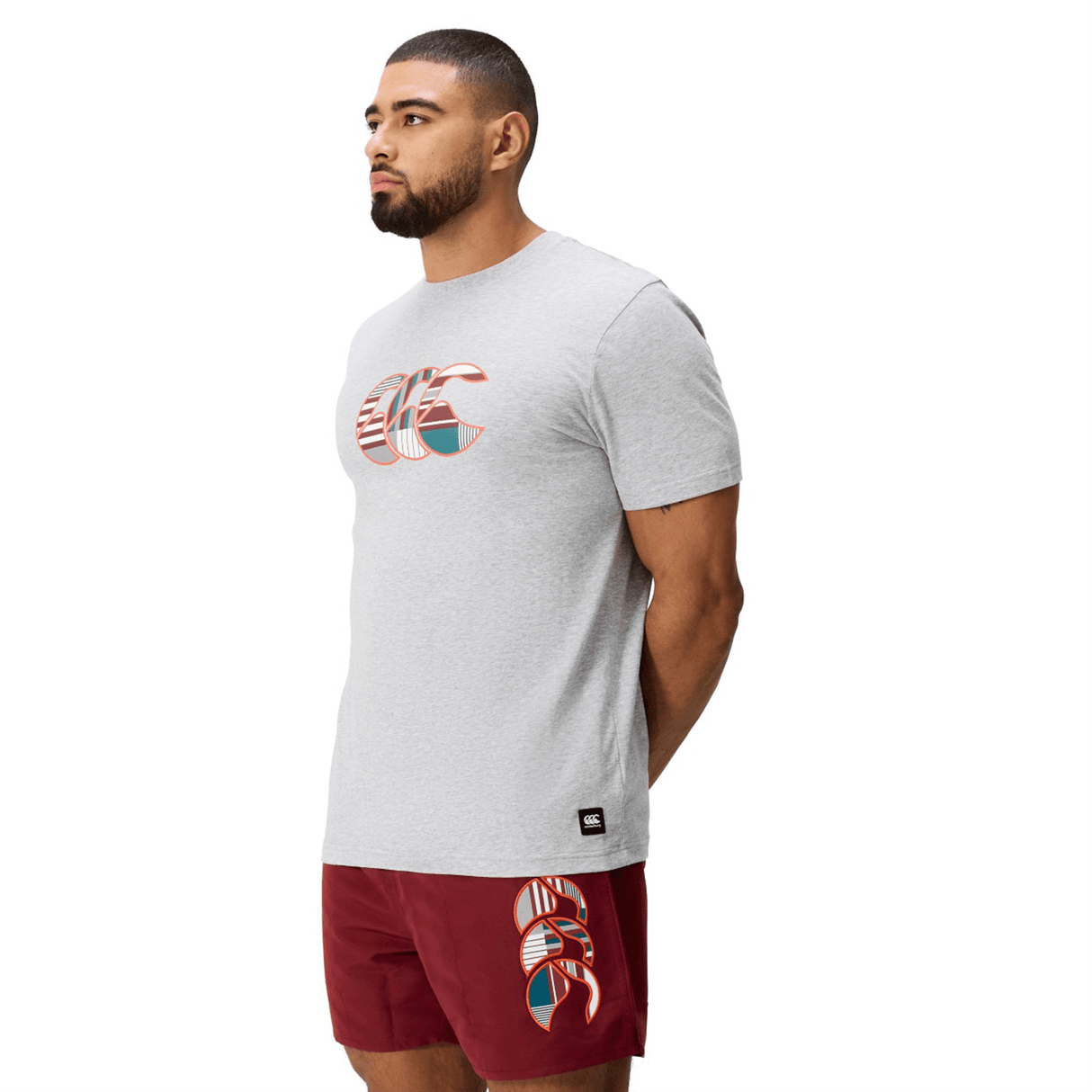 A man faces left, wearing a light gray Canterbury Uglies Logo T-shirt and red shorts with a matching graphic design, set against a plain white background.