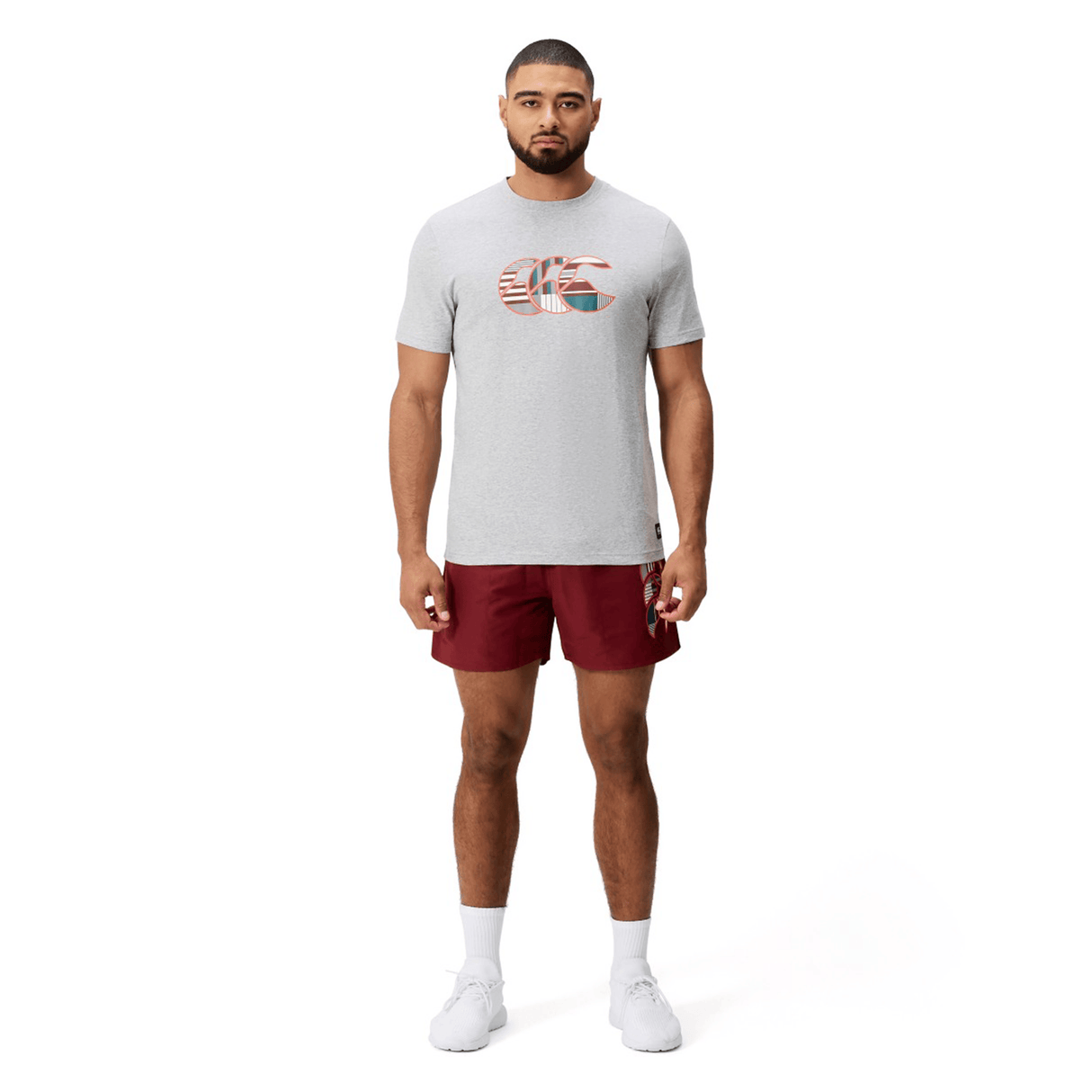 A man faces forward wearing a Canterbury Uglies Logo T-shirt by Canterbury, featuring a UK flag graphic, paired with maroon shorts, white socks, and white athletic shoes on a plain white background.