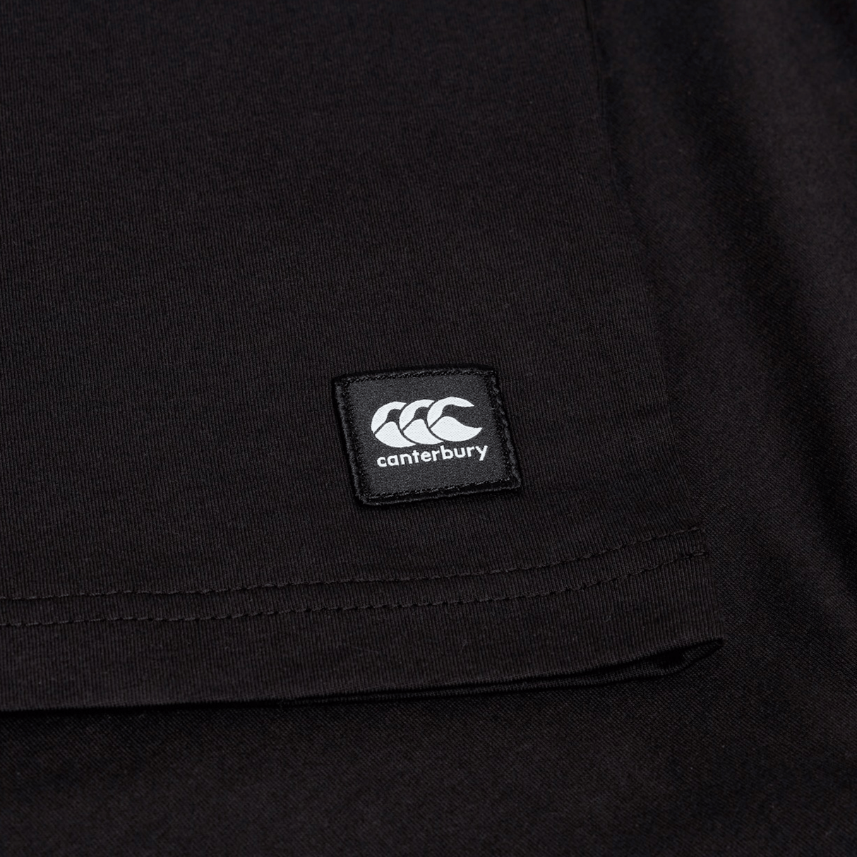 Close-up of the Canterbury Uglies Logo T-shirt in a black cotton blend, featuring a stitched rectangular Canterbury logo patch near the hem, inspired by classic Canterbury rugby shirts.