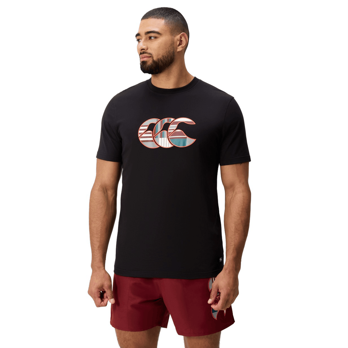 A bearded man wears a black Canterbury Uglies Logo T-shirt—a cotton blend tee with a patterned logo—and maroon athletic shorts, standing against a white background.
