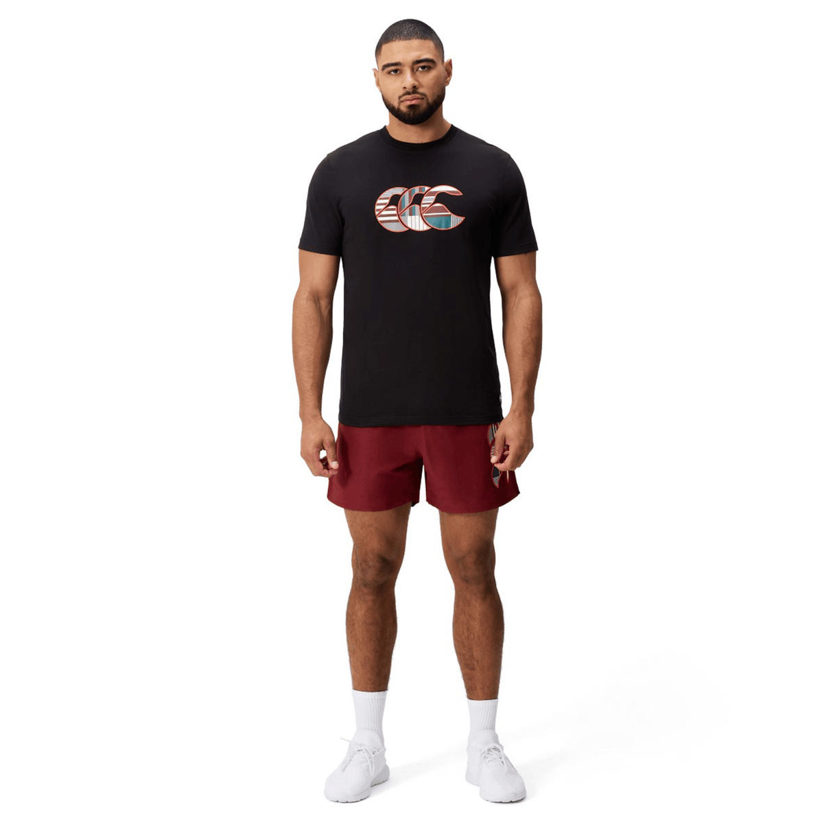 A man faces forward against a plain white background, wearing a black Canterbury Uglies Logo T-shirt with a patterned logo, maroon shorts, white socks, and white athletic shoes.