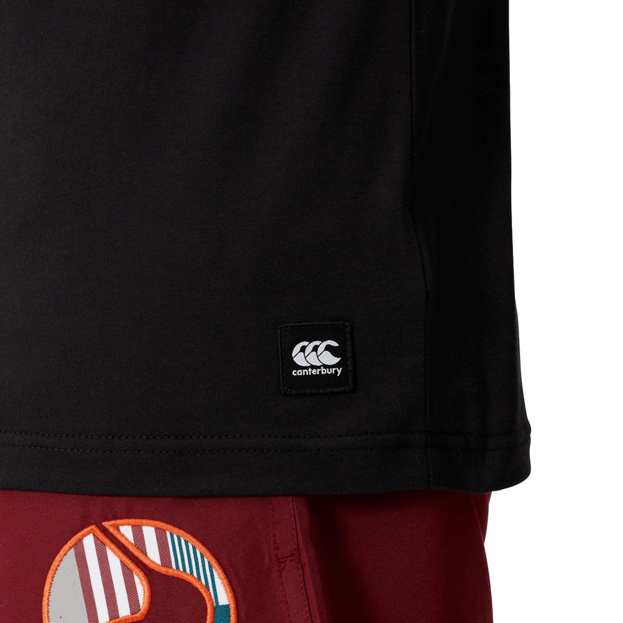 Close-up of the Canterbury Uglies Logo T-shirt—a black cotton-blend tee with the Canterbury logo tag—paired with red shorts featuring a vibrant abstract pattern.