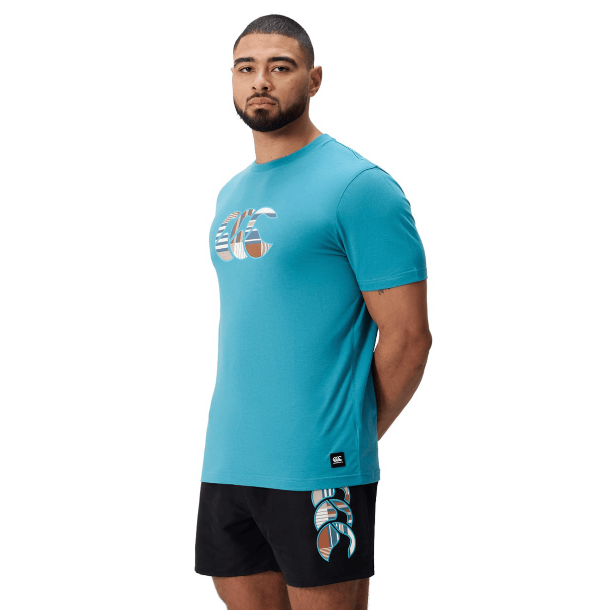 A man faces forward, wearing the Canterbury Uglies Logo T-shirt by Canterbury with a geometric graphic, paired with black shorts featuring a matching design on the left leg, set against a plain white background.
